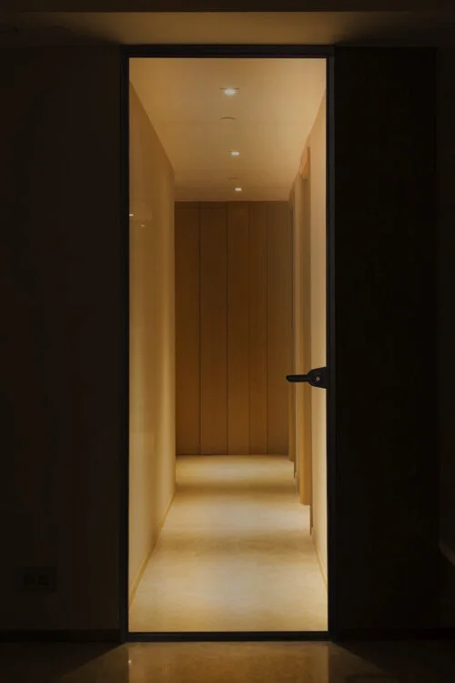 View through an open doorway into a well-lit, narrow corridor with wooden wall panels and ceiling lights.