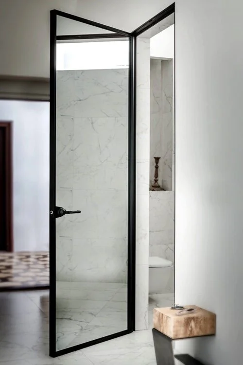 Open black-framed glass door leading to a modern bathroom with white marble walls and a toilet, with a small wooden stool beside it.