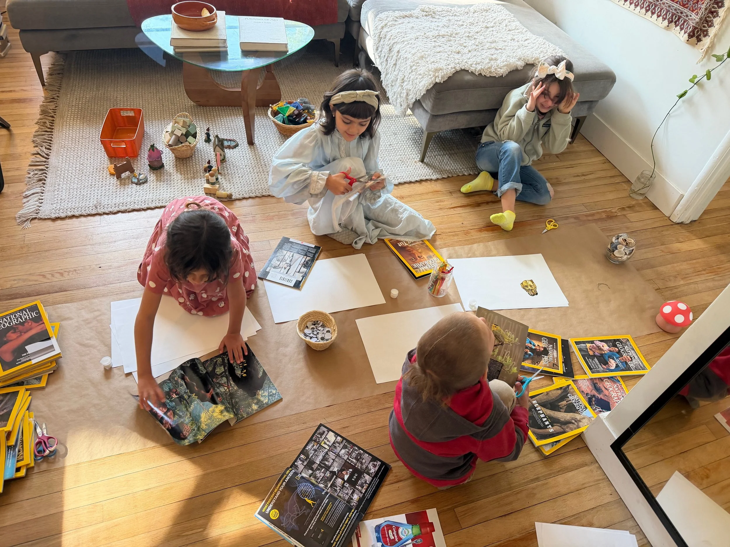 Four children sitting on the wooden floor engaging in arts and crafts activities. One girl is reaching for a book, another girl in a white outfit is cutting paper, a boy is flipping through a magazine, and a girl in a gray hoodie is holding her head. There are scattered magazines, books, scissors, paper, and small craft supplies around them. The room has a rug, a glass coffee table, and a gray couch in the background.