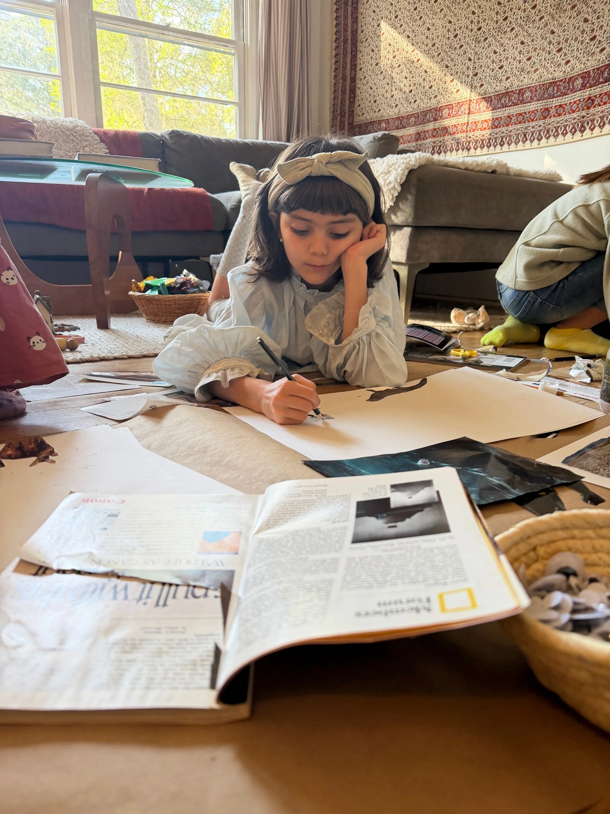 A young girl with dark hair and a beige headband is lying on the floor, drawing with a pen on a large sheet of paper. The floor is cluttered with magazines, papers, and art supplies. In the background, there is a sofa, a window with lush green trees outside, and a wall hanging with a red and beige pattern.