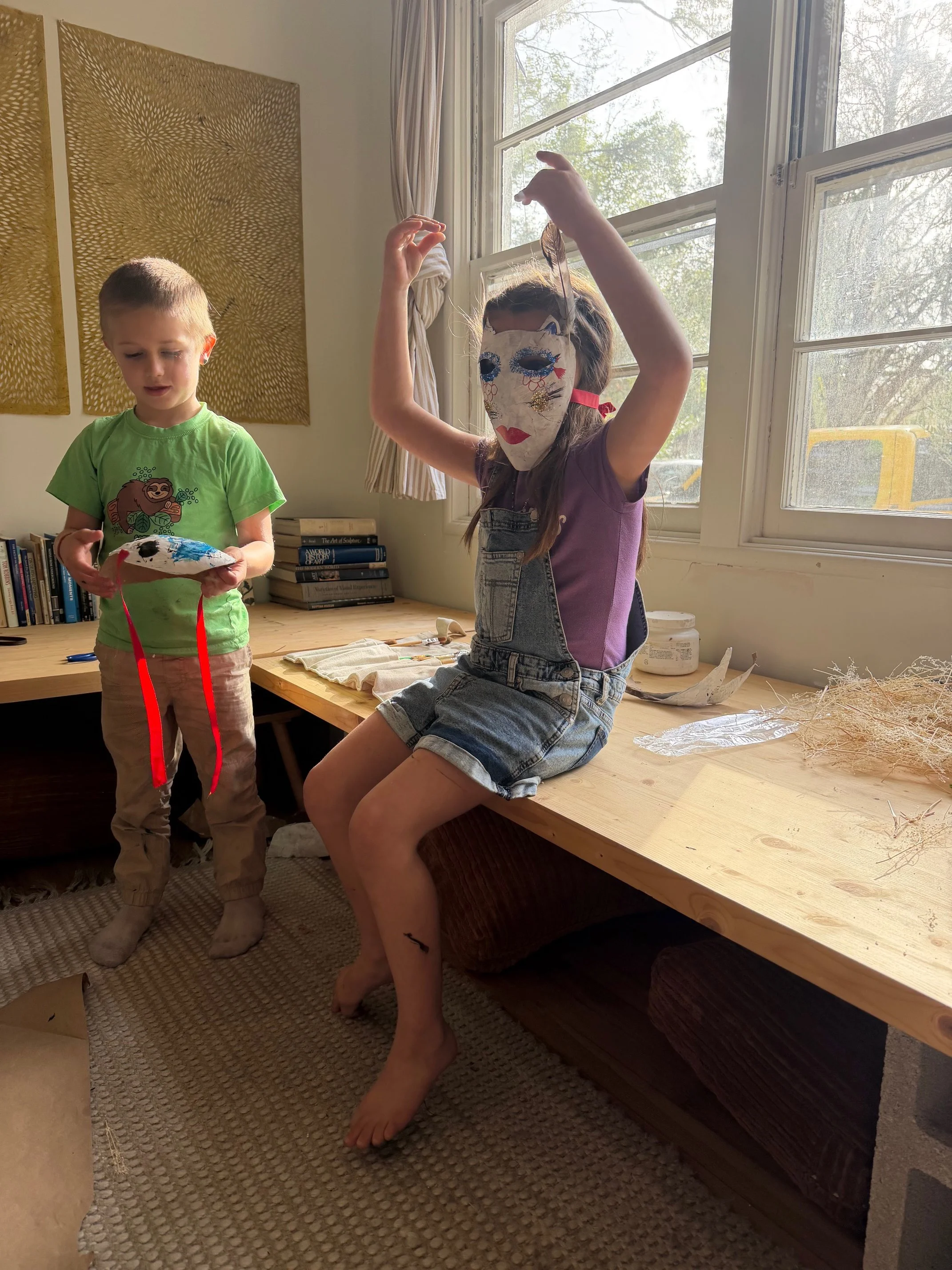 Two children, a boy and a girl, are in a room with a wooden desk and large window. The girl is sitting barefoot on the desk wearing a mask with painted and glued features, holding a string attached to a mask. The boy is standing next to her, holding a decorated mask. The room has books on the desk, art supplies, and a decorative yellow wall hanging.