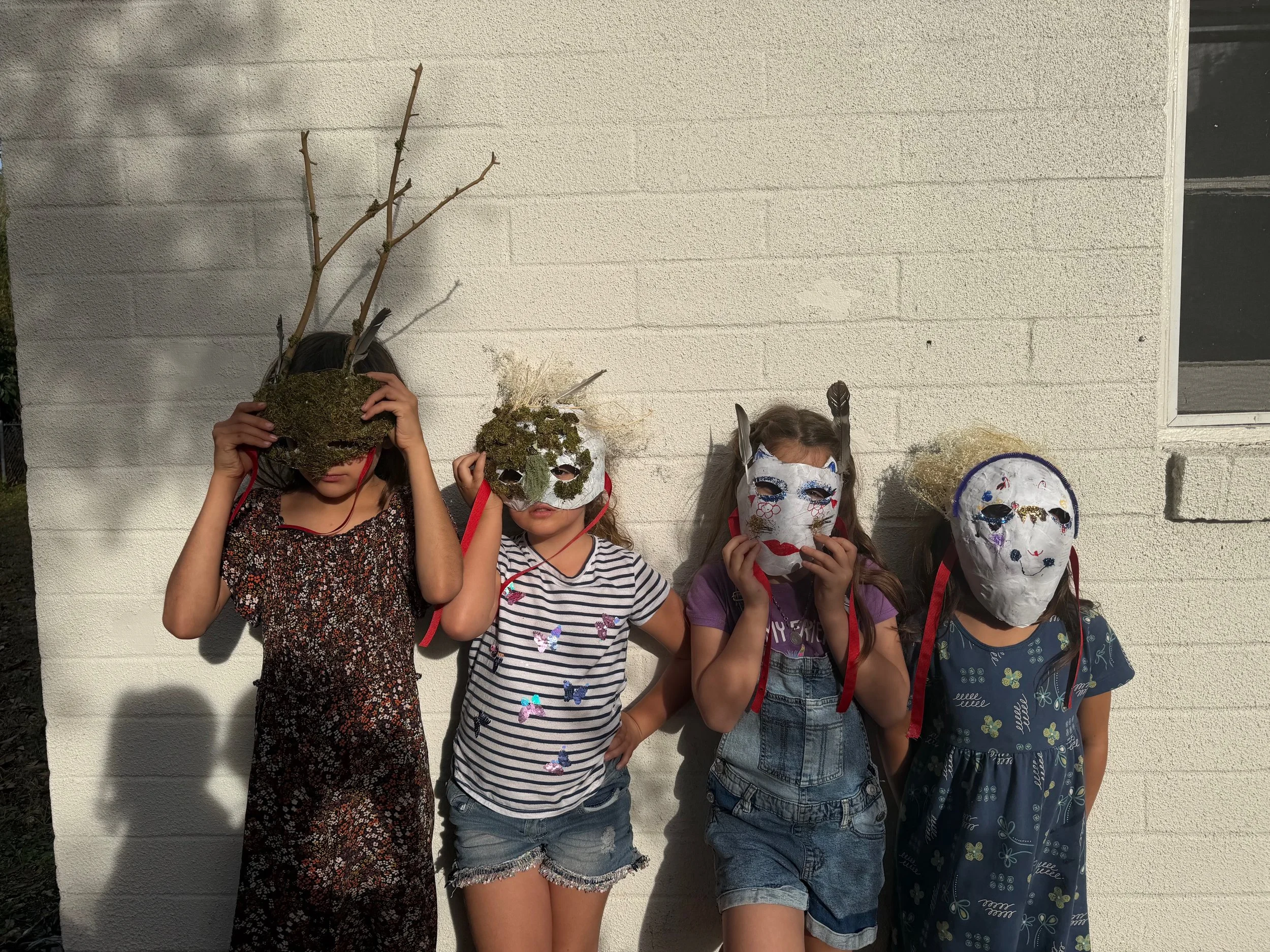 Four children wearing handcrafted masks and standing outdoors against a white brick wall, with one child in a black dress with flowers, another in a striped shirt and denim shorts, the third in a purple shirt and denim overalls, and the fourth in a blue dress with yellow flowers.