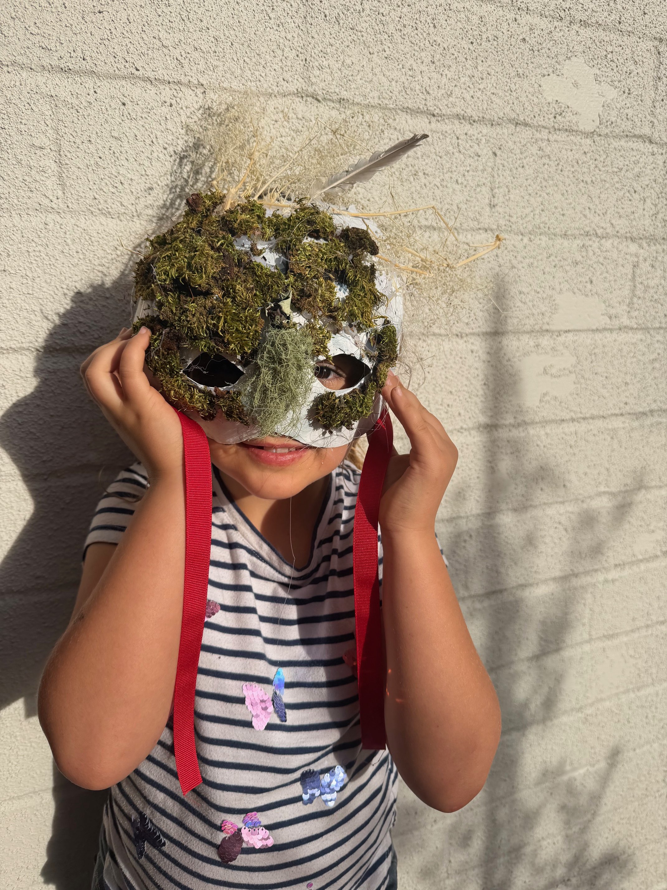 A child holding a white mask decorated with moss, found objects, and feathers, standing against a textured white wall.