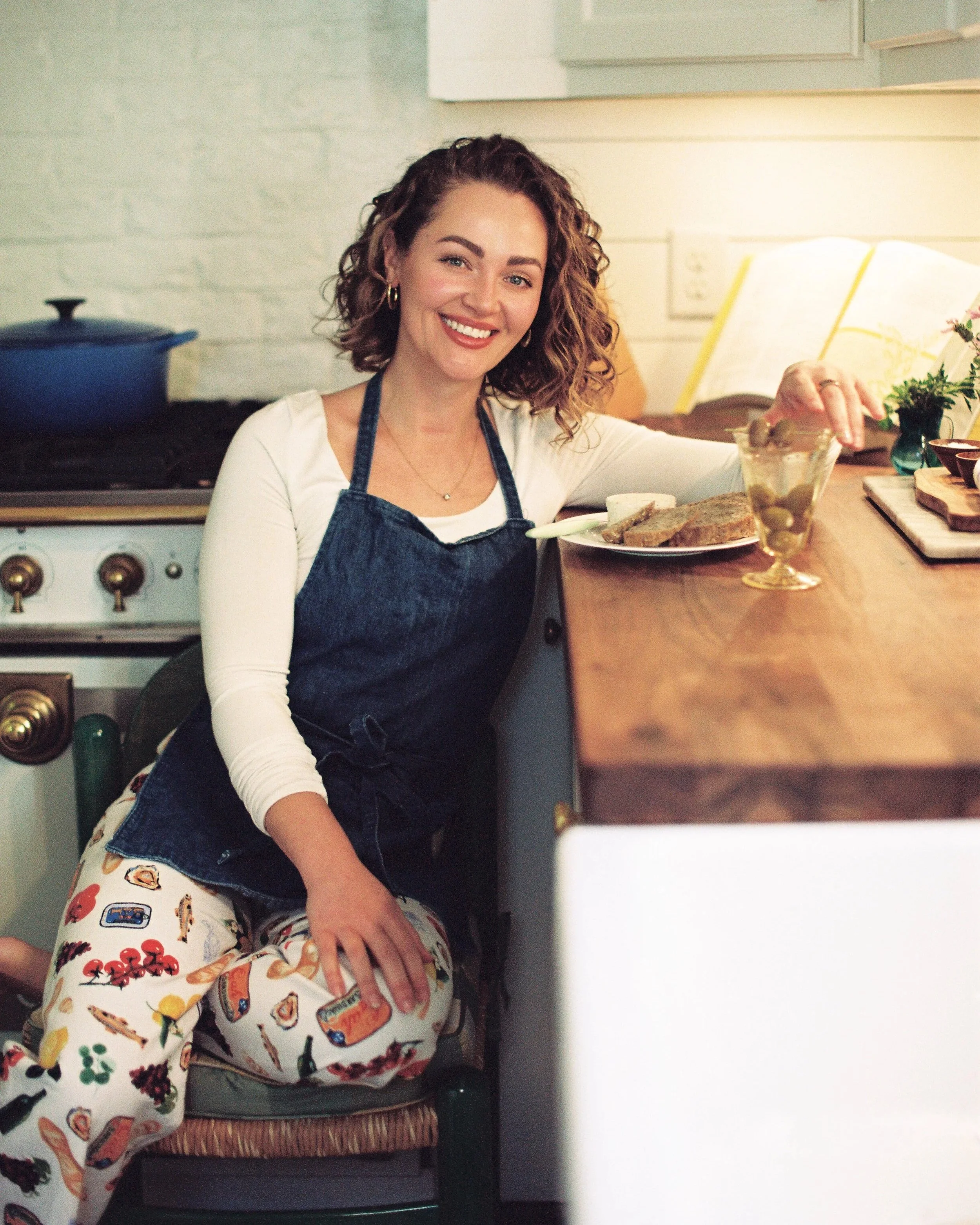 A woman with curly hair smiling while sitting in a kitchen. She is wearing a white long-sleeve shirt and a dark apron. She is holding a plate of food and has a glass dish in front of her. The background includes a stove, kitchen cabinets, and kitchen accessories.