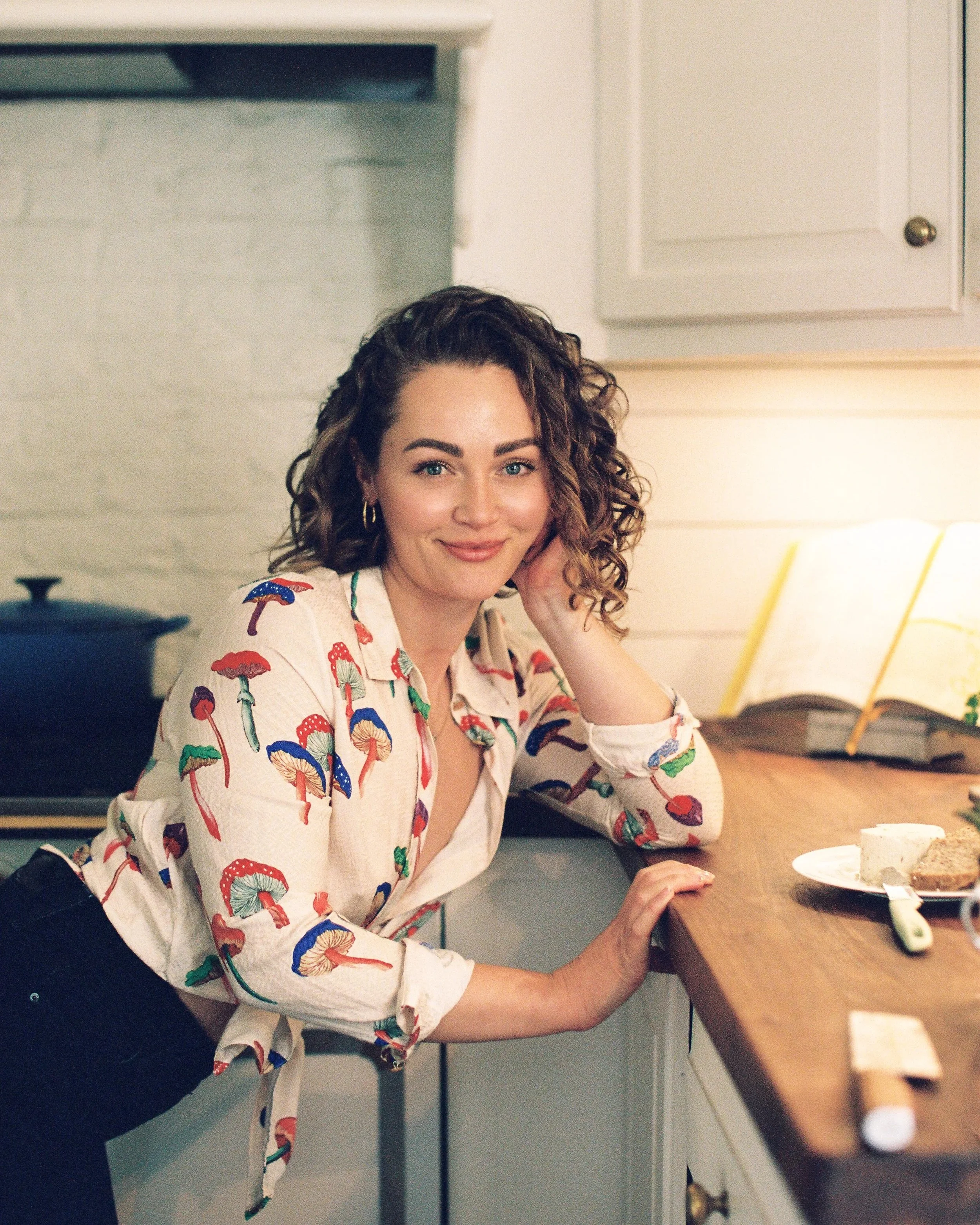 A woman with curly brown hair, blue eyes, and hoop earrings is seated at a wooden table in a kitchen, looking at the camera with a smile. She is wearing a white shirt with a colorful mushroom pattern, and has her left hand resting on the table near a plate with cake. There is a yellow-striped paper straw in a yellow beverage cup on the table. In the background, there are white cabinets and a section of a white brick wall.