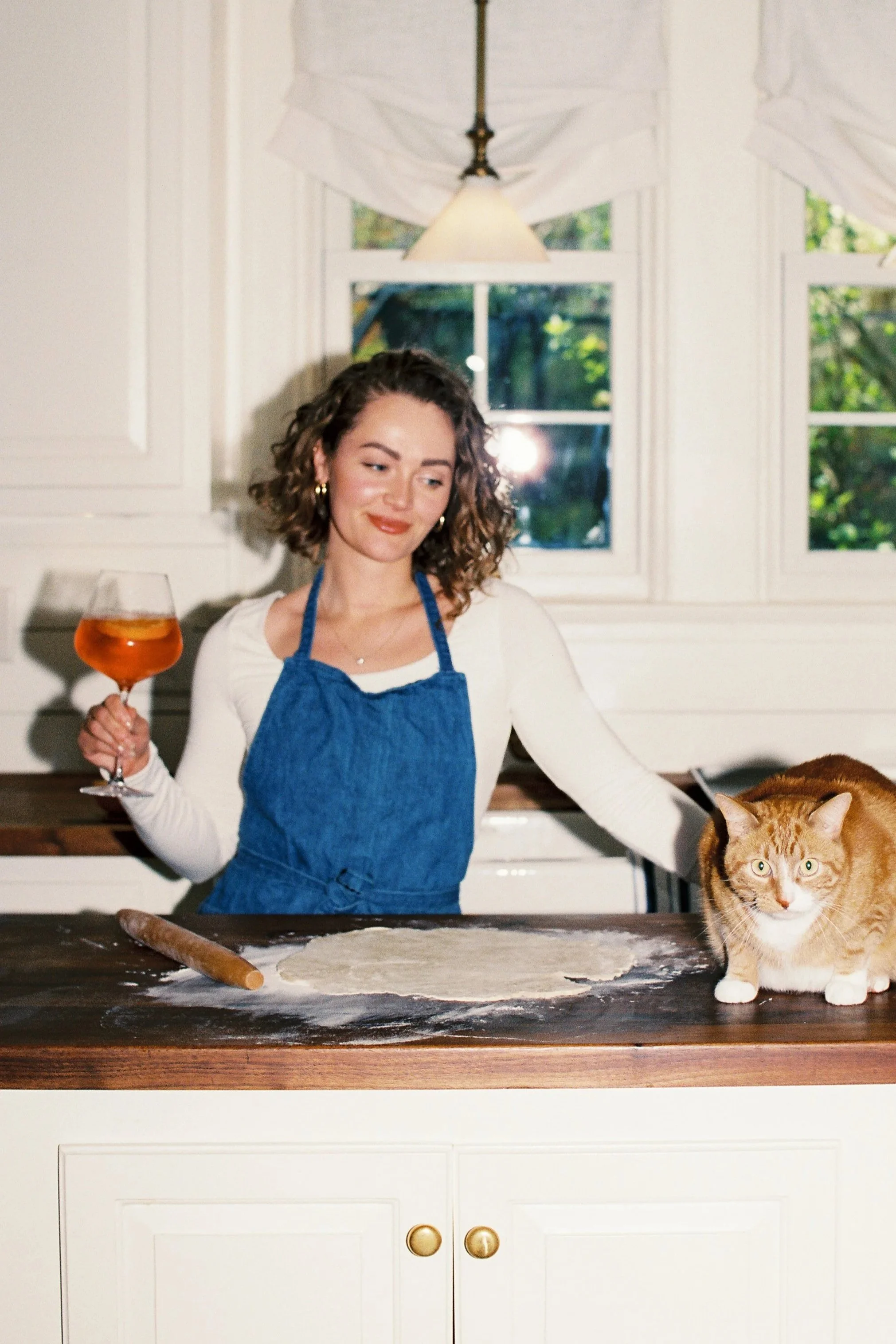 A woman with curly hair wearing a white shirt and blue apron stands in a kitchen, holding a glass of wine in her right hand and looking at a ginger cat on the counter. The counter has a rolling pin and some dough. Behind her is a window with white curtains and a hanging lamp.