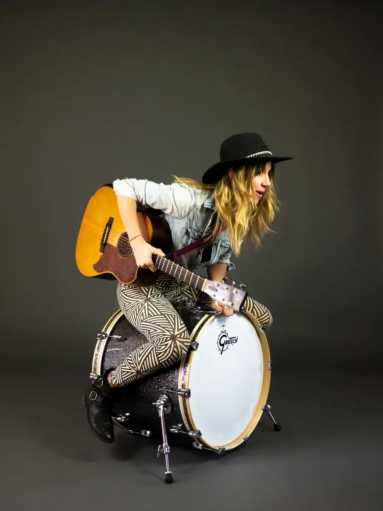 Tiffany Christopher is playing an acoustic guitar while sitting on a bass drum against a dark background. She will perform at The StormHold on November 22, 2026.