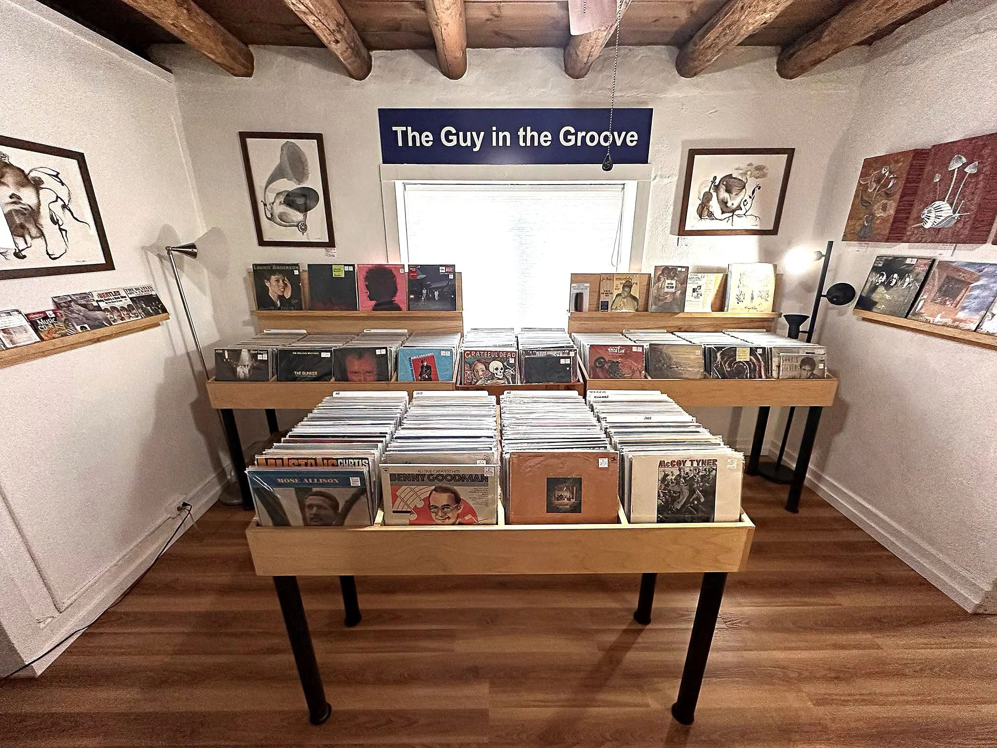 The Guy  in the Groove record store with wooden display shelves has a cozy, rustic ambiance with wooden ceiling beams and hardwood floors.The walls feature art by Linda Storm.