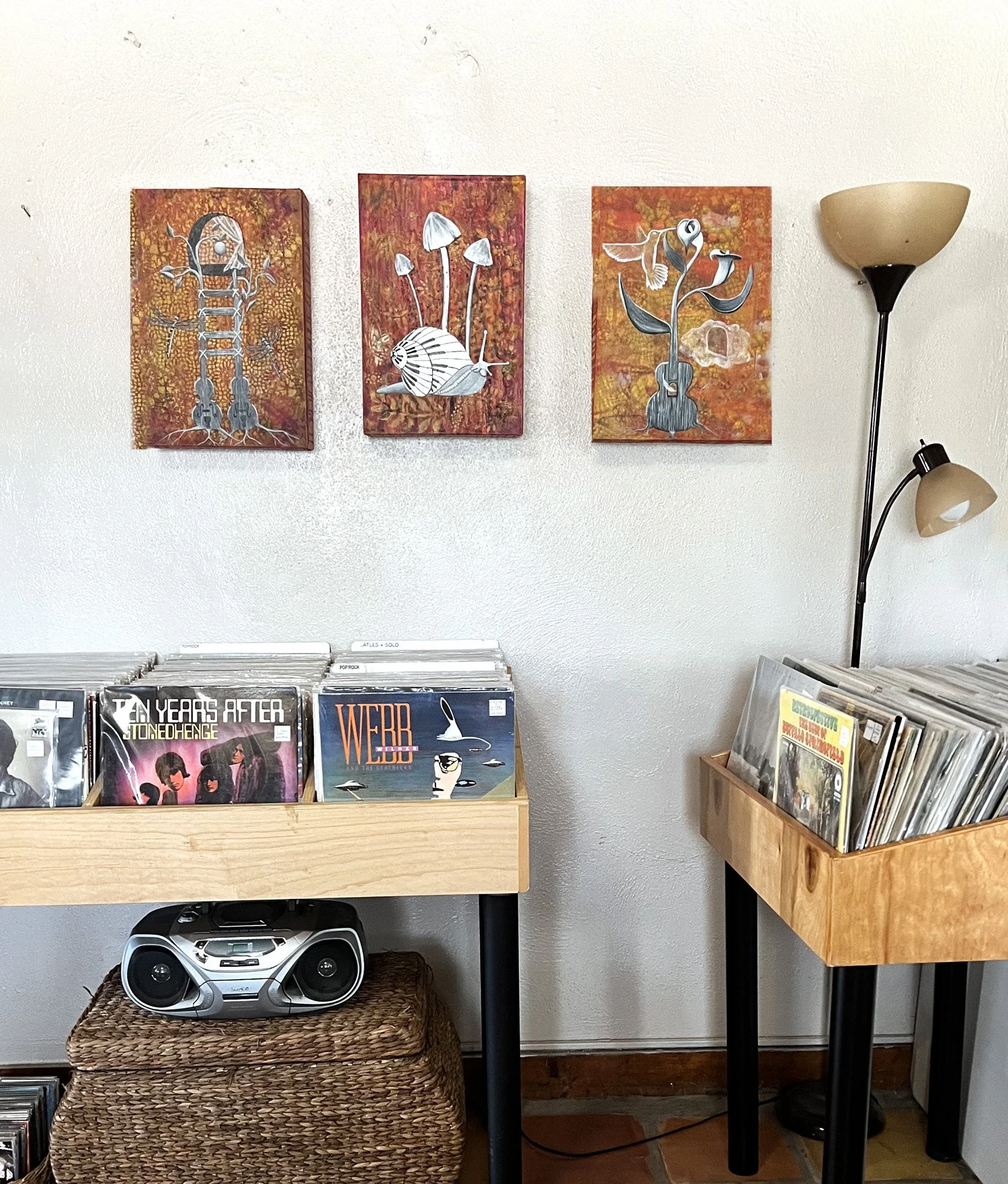 Interior of a record store with three abstract paintings on the wall, records in wooden bins, a stereo system on a woven basket, and a dual-headed floor lamp.