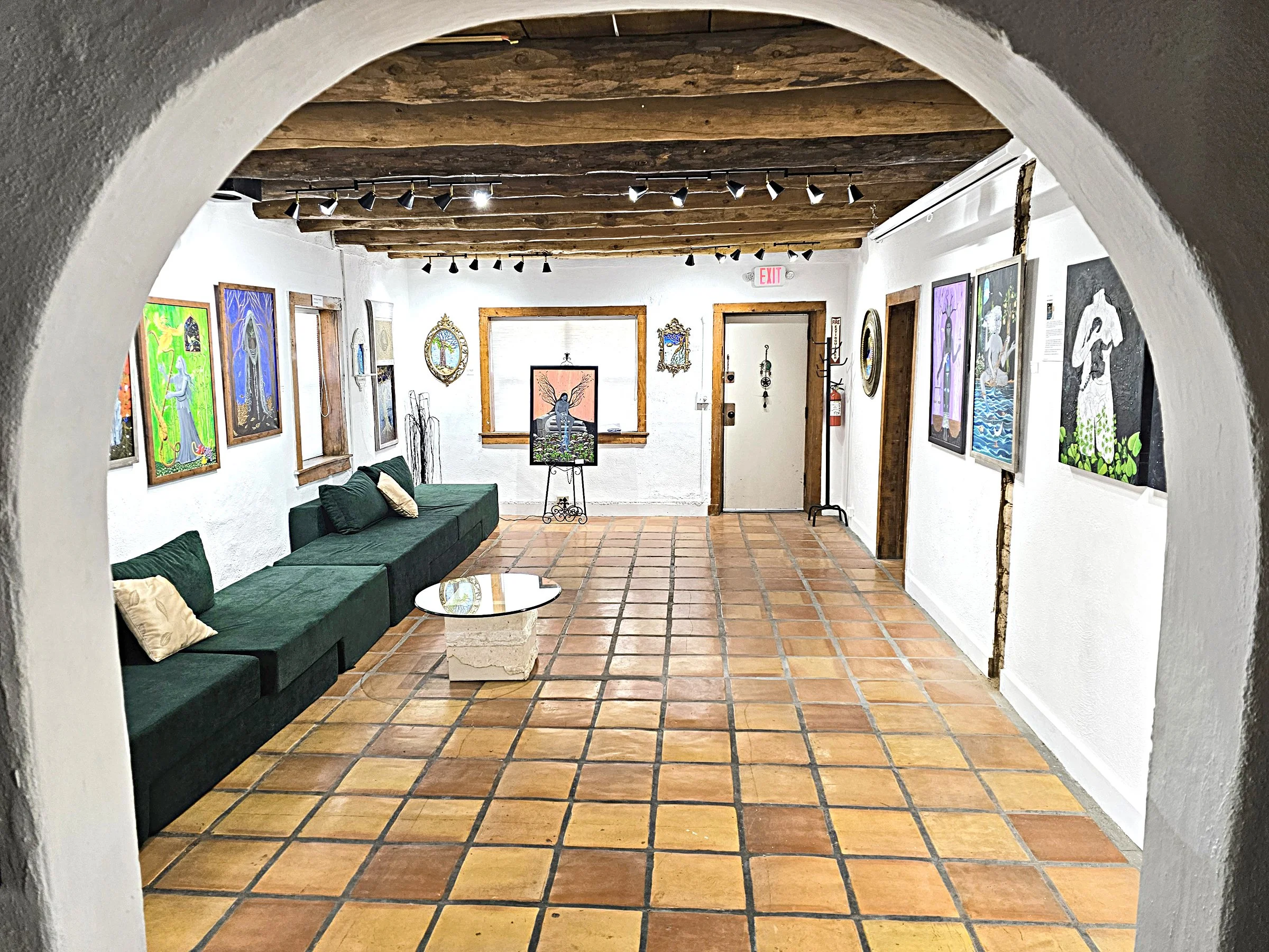 An art gallery with white walls, wooden ceiling beams, and terracotta tile floors. Several colorful paintings by Linda Storm are displayed on the walls.