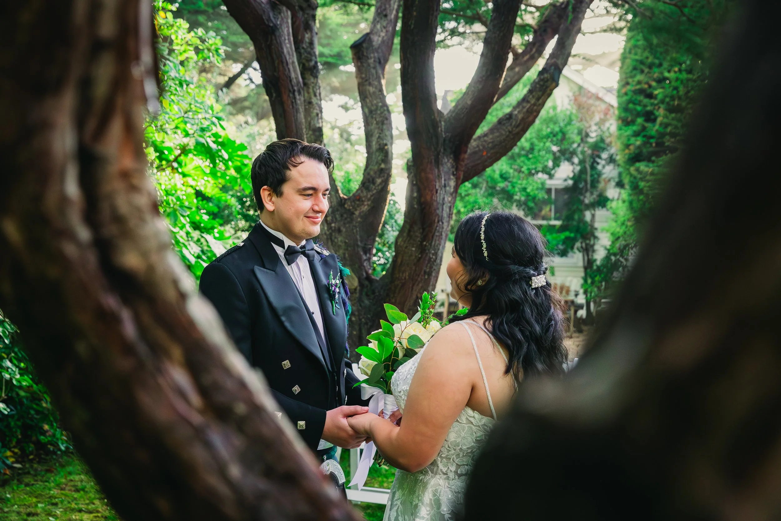 A bride and groom stand face-to-face, holding hands, during their wedding ceremony outdoors among trees, with the groom dressed in a formal black tuxedo and the bride in a lace white wedding dress holding a bouquet of white flowers and green leaves.