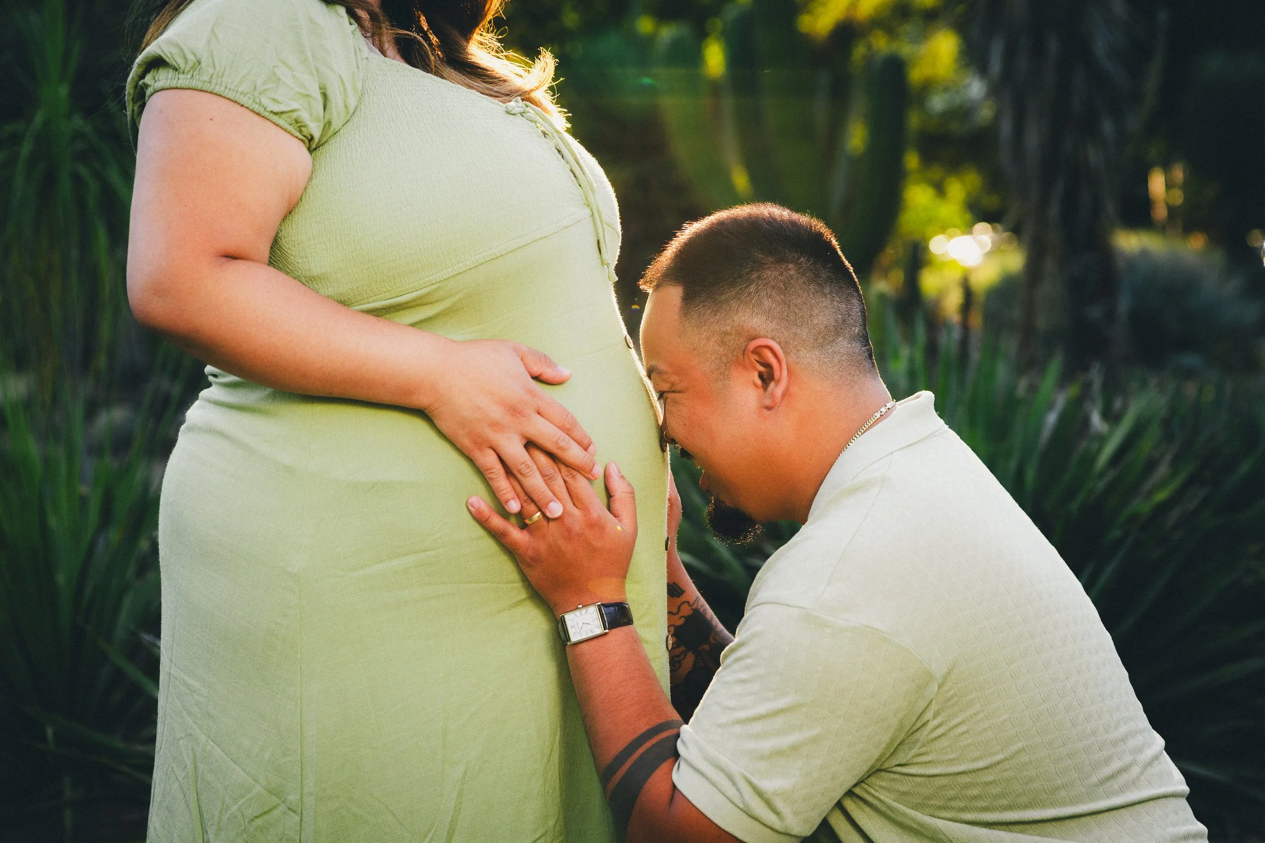 A man kneeling and kissing a pregnant woman's belly outdoors during sunset, with both wearing light-colored clothing and lush greenery in the background.