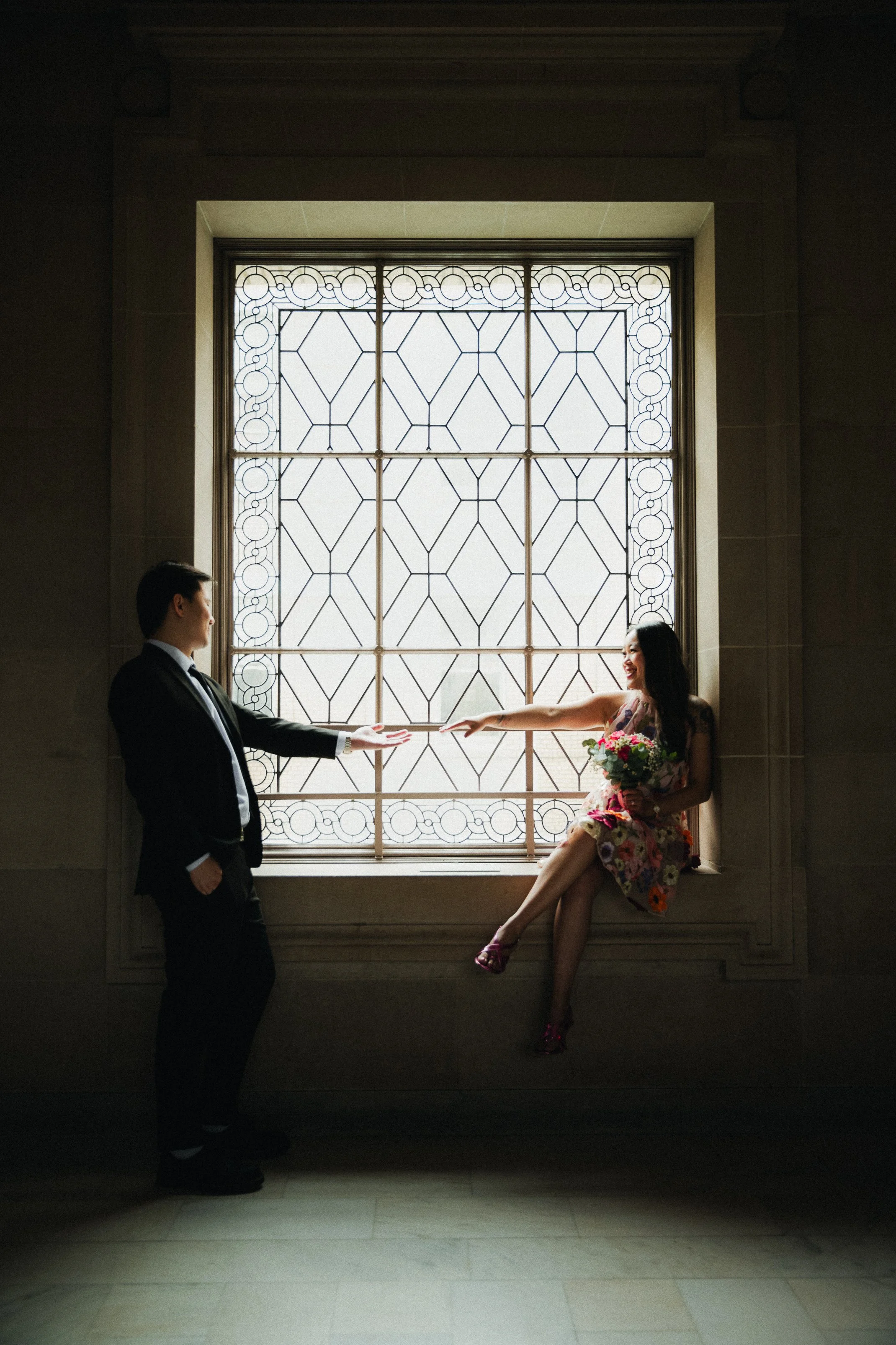 A man in a suit and a woman in a colorful dress exchanging a bouquet of flowers in front of a large decorative window.