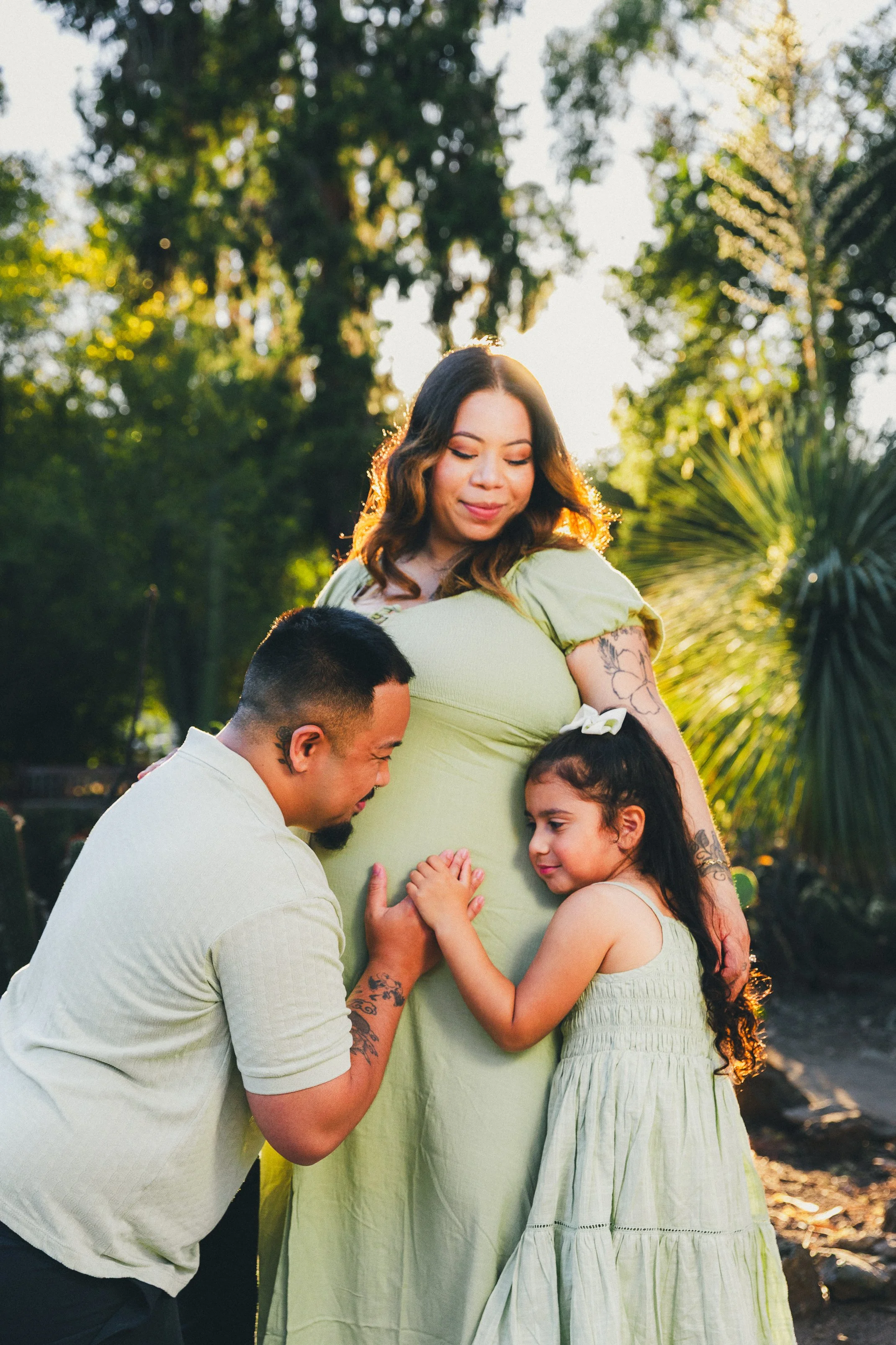 A couple and a young girl gather around a pregnant woman outdoors during sunset, affectionately touching her belly, with lush green trees in the background.