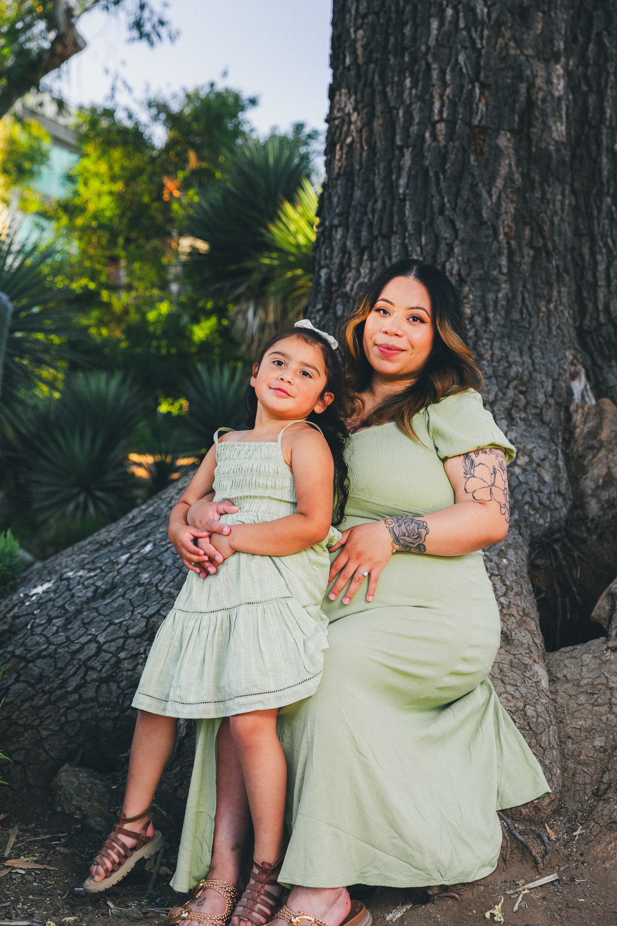 A woman and young girl posing outdoors in front of a large tree. Both wearing matching light green dresses. The woman has tattoos on her arm and shoulder. They are sitting on a fallen branch with greenery in the background.