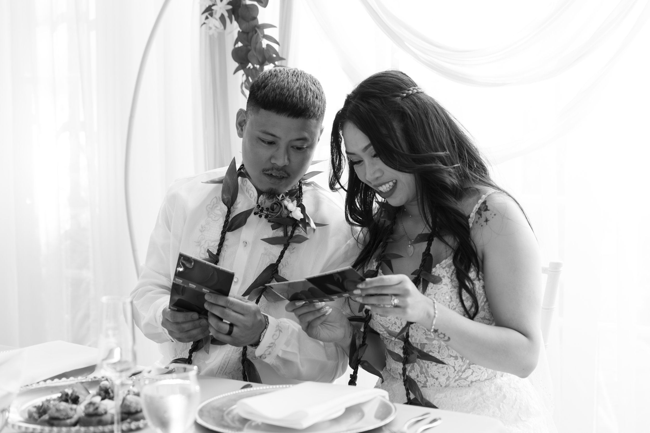 A couple in wedding attire looking at photographs during a wedding celebration, with a table of food and drinks in front of them.