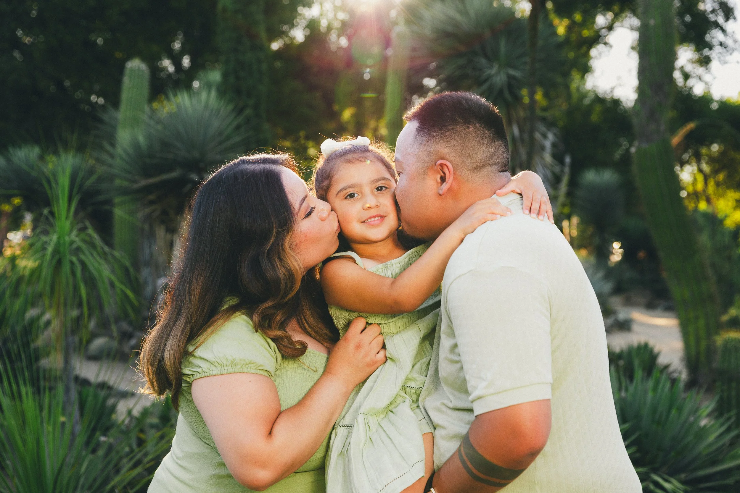 A family of three, a woman, a man, and a young girl, sharing a kiss outdoors with greenery and cactus plants in the background, during sunset.