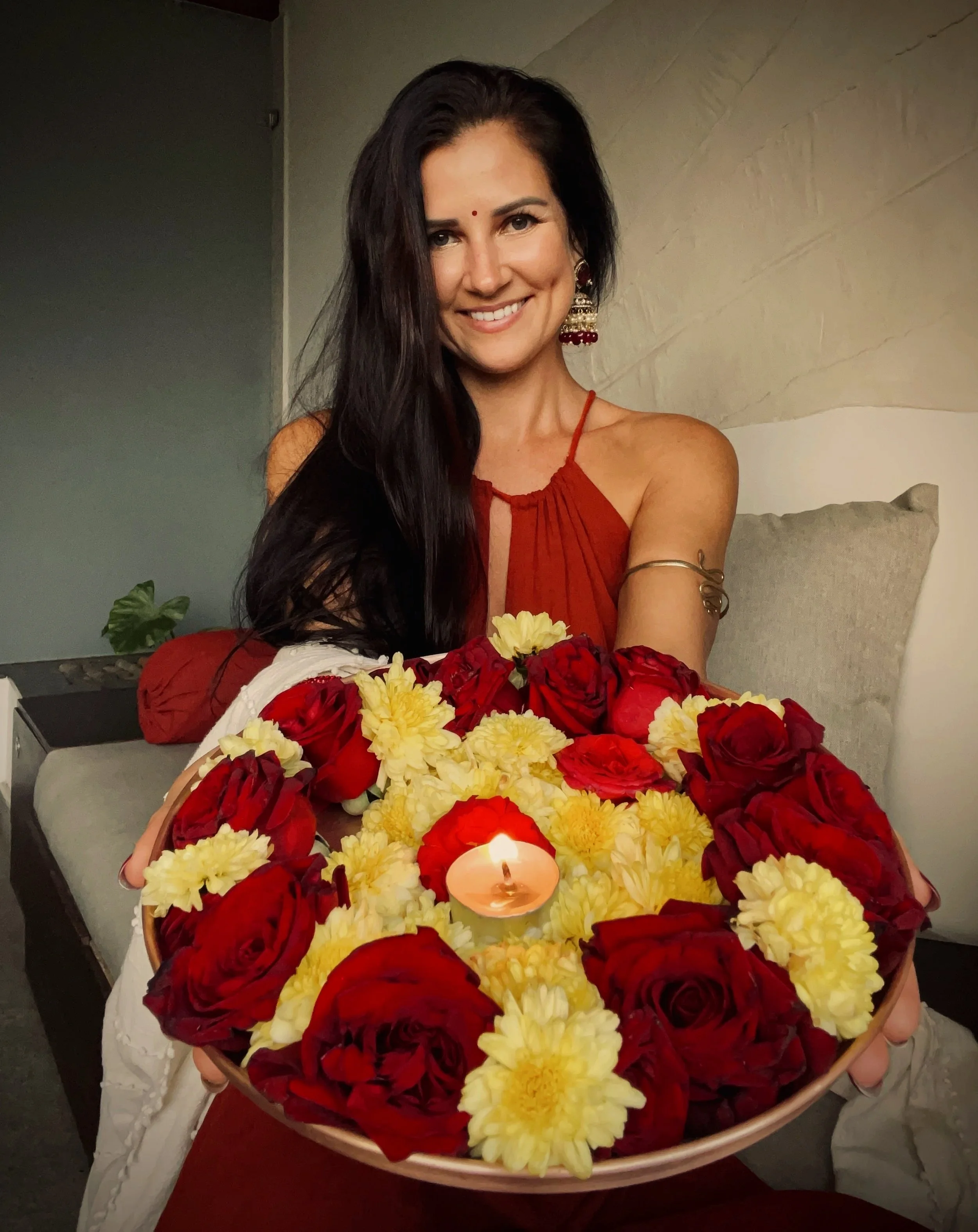 A woman with long dark hair, wearing traditional Indian earrings and a red dress, smiling and holding a large tray of red roses, yellow chrysanthemum flowers, and a lit candle in the center.