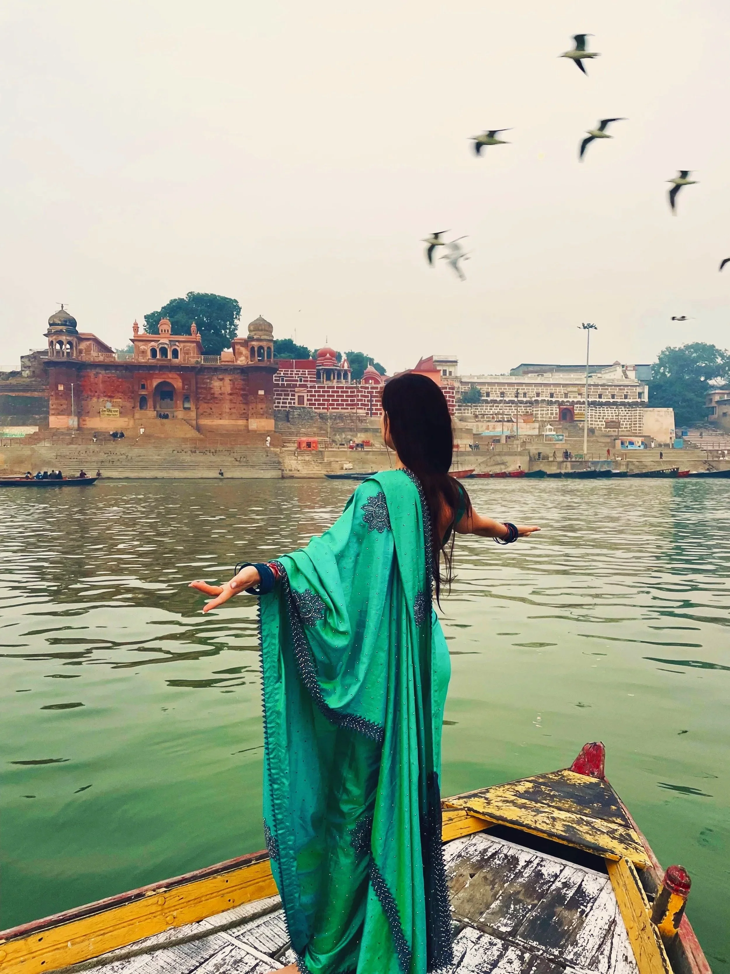 A woman in a turquoise saree standing on a boat with arms outstretched on a river, with historical red sandstone building and white fort on the opposite bank, and birds flying overhead.