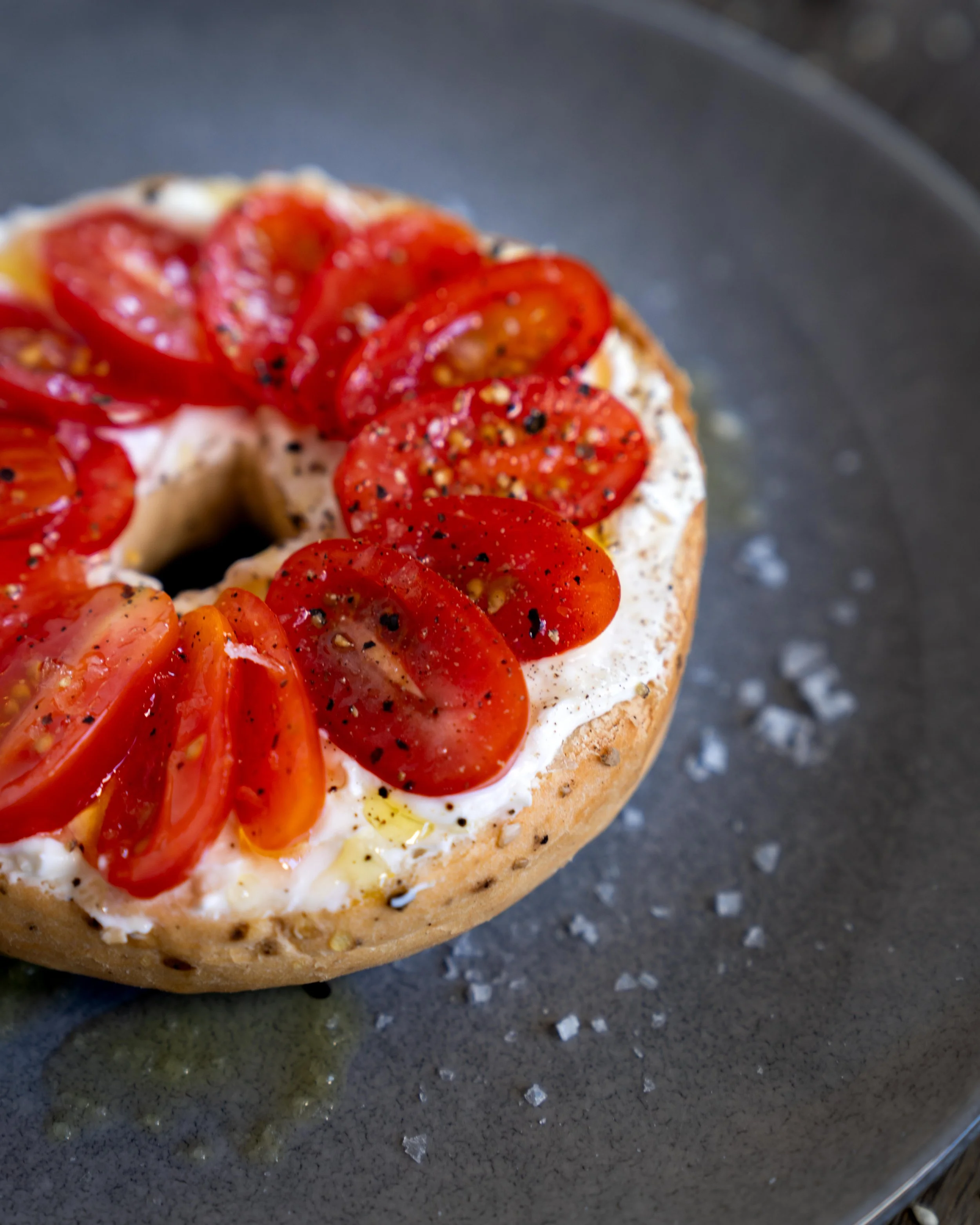 Close-up of a baked bagel topped with cream cheese, cherry tomato slices, black pepper, served on a black plate.