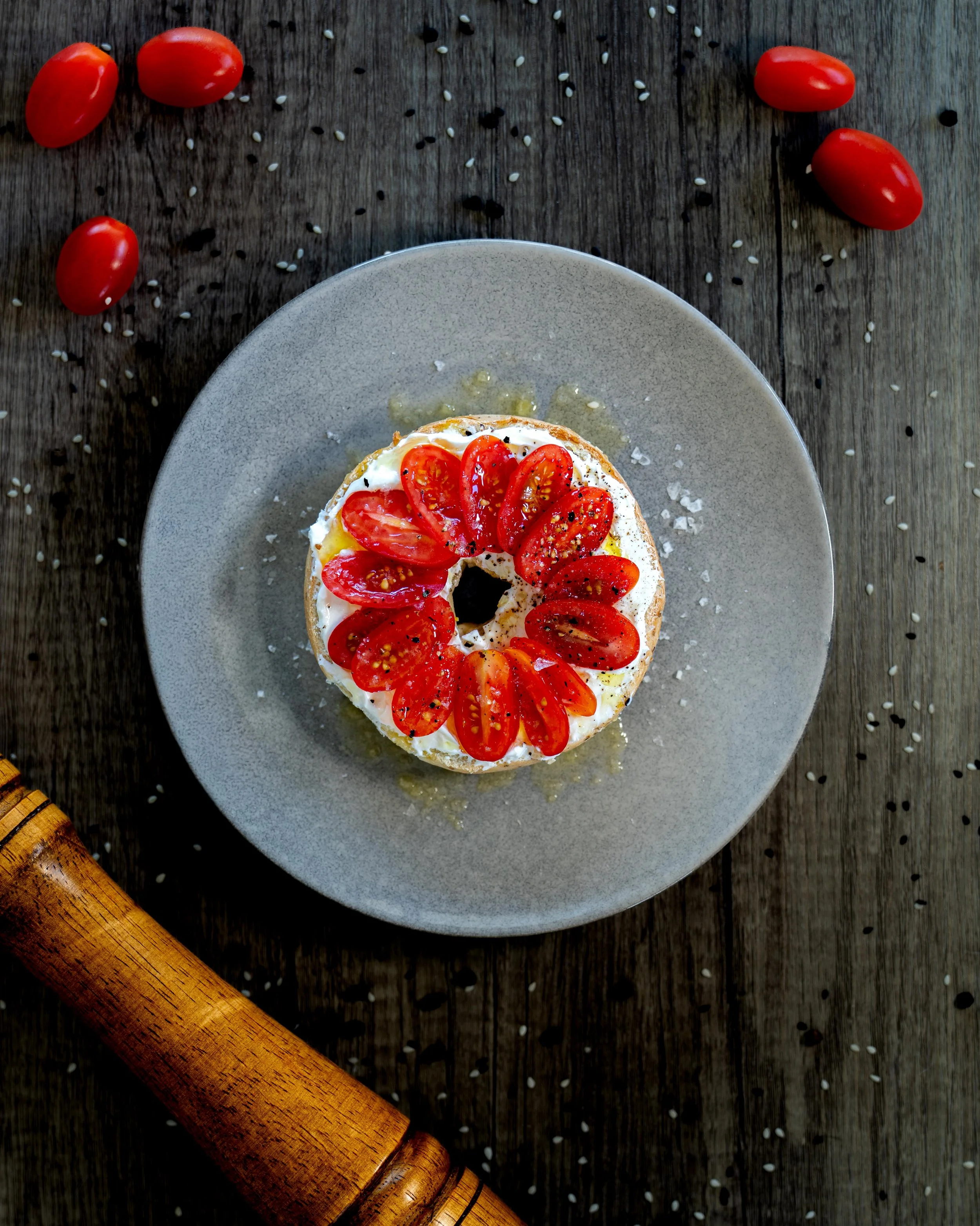A plate with a cake topped with cherry tomatoes and black pepper on a dark wooden surface, with additional cherry tomatoes and sesame seeds scattered around.