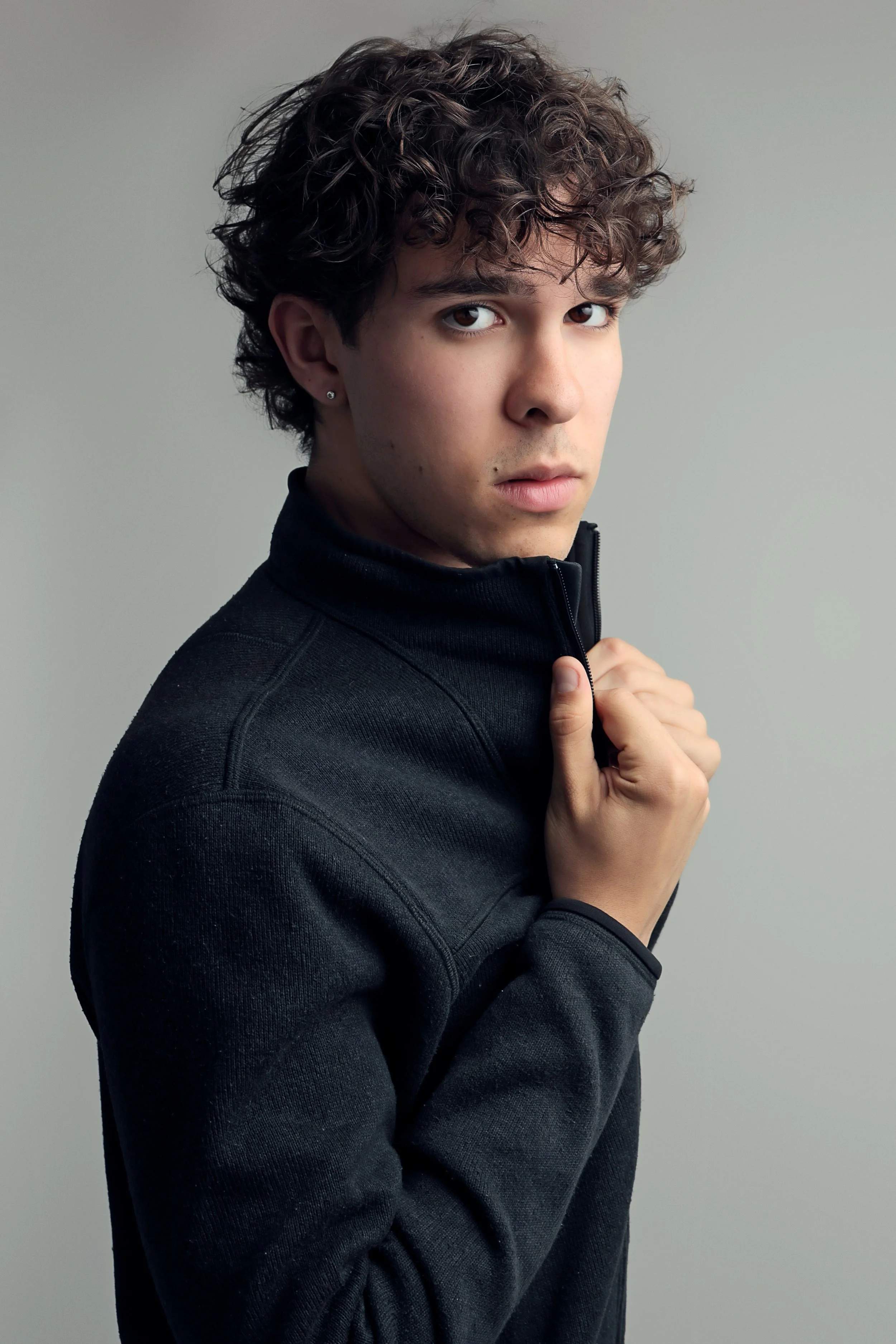 A young person with curly dark hair wearing a black jacket holding the zipper with one hand, looking into the camera against a plain light gray background.
