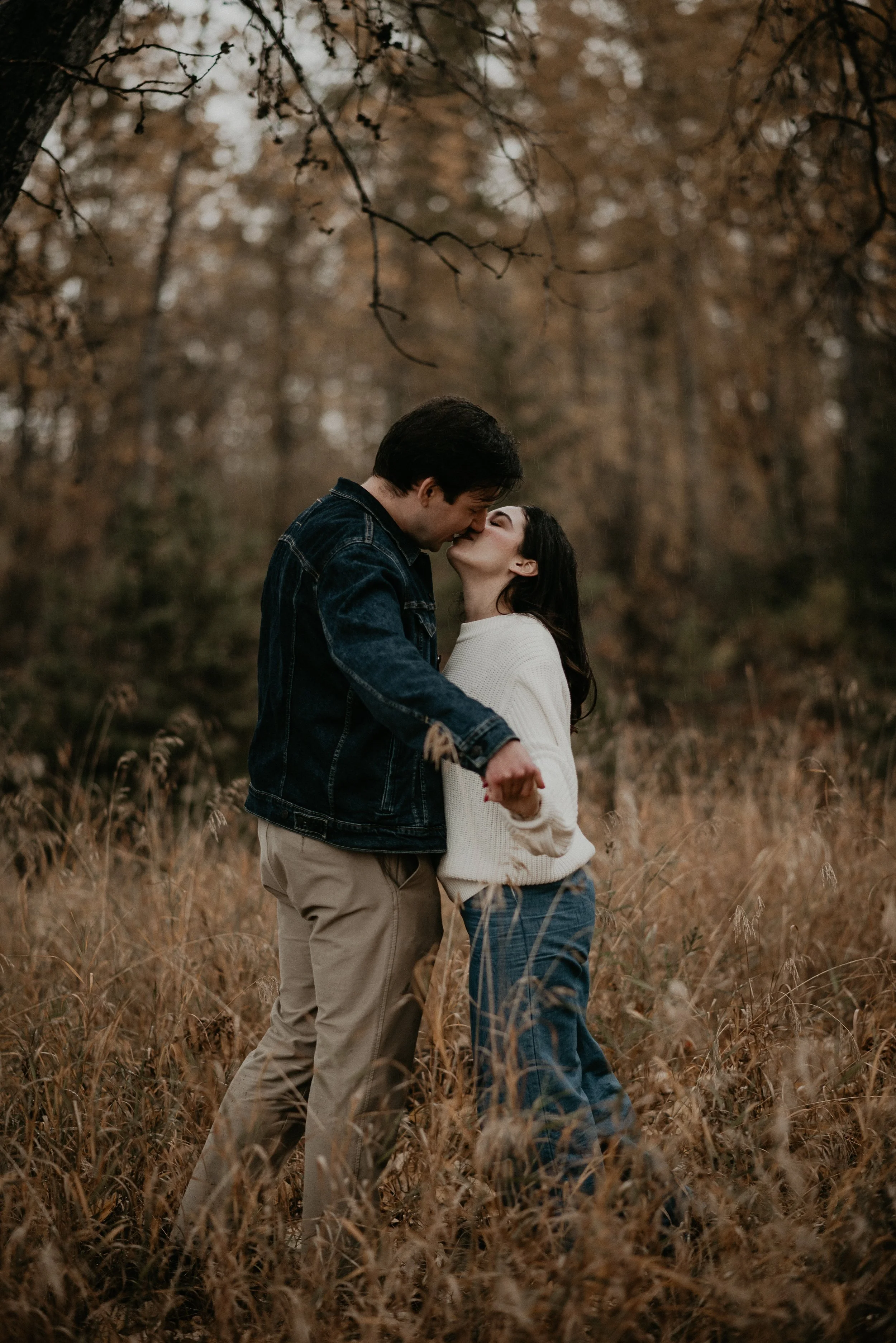 Couple sharing a kiss in a grassy field during their engagement session at McKenzie Trails in Red Deer, Alberta, photographed by Danielle Aichelle Photography