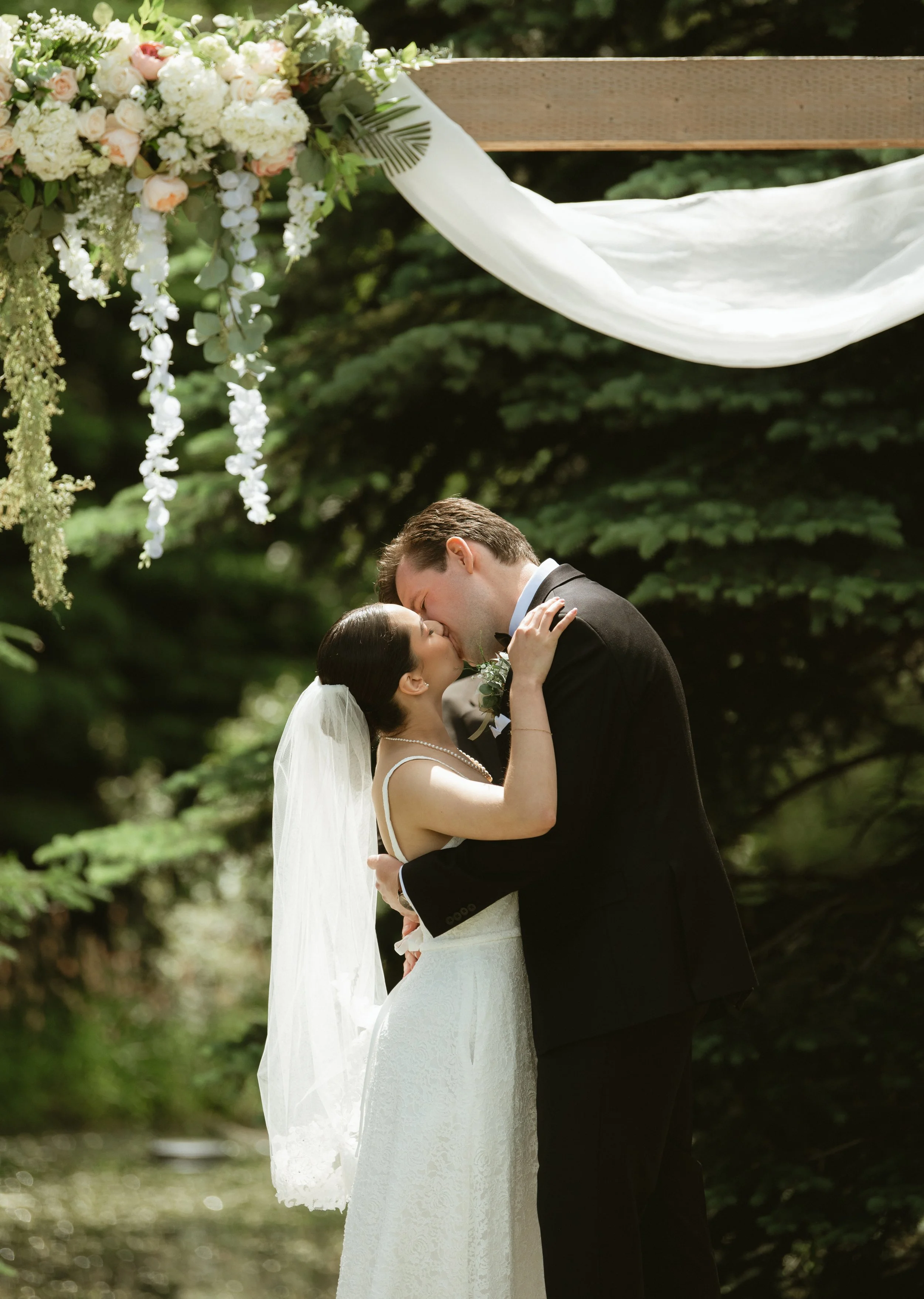 Bride and groom sharing their first kiss beneath the floral wedding arbor during their ceremony at Pine & Pond Wedding Venue in Ponoka County, Alberta, photographed by Danielle Aichelle Photography