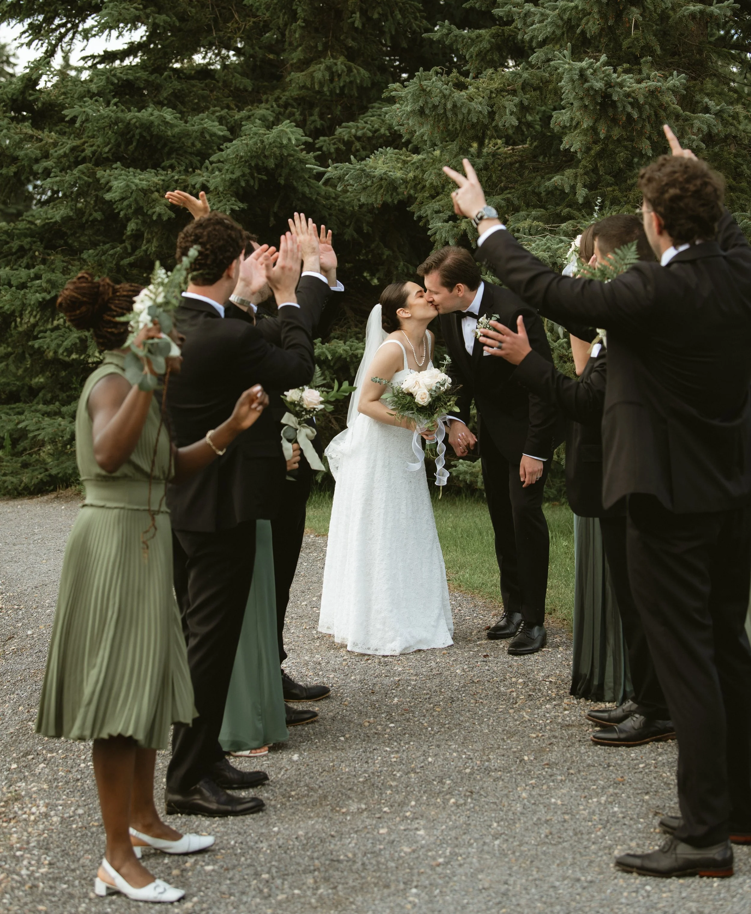 Wedding ceremony with a bride and groom kissing, surrounded by wedding guests dressed in formal attire, outdoors near a large green pine tree.