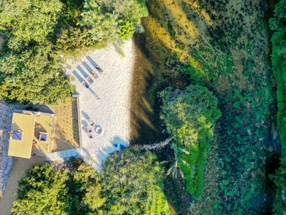 An aerial view of a sandy beach with lounge chairs, a house with a yellow roof, surrounded by green trees, water, and vegetation.