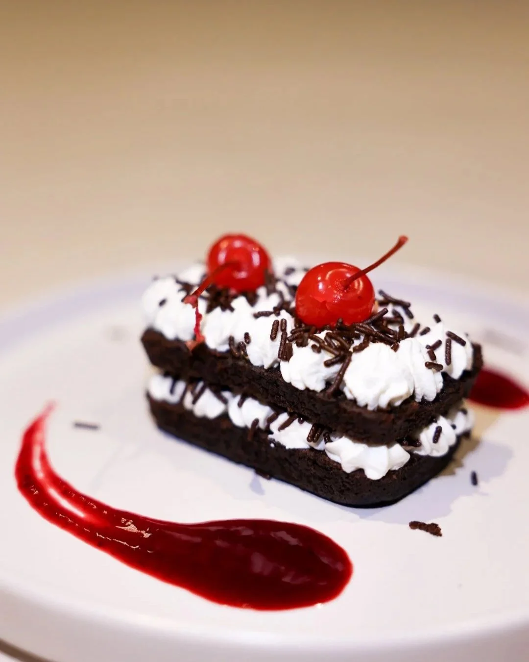 Chocolate layered cake with whipped cream, chocolate sprinkles, cherries, and red sauce on white plate