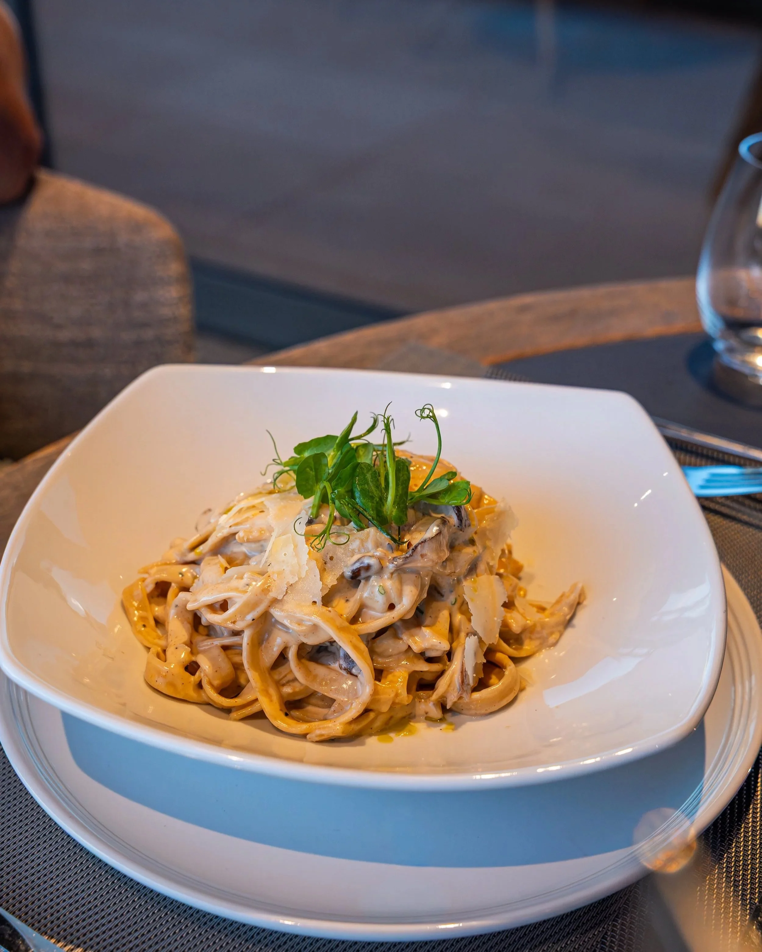 A white bowl of creamy mushroom pasta garnished with microgreens, on a table with a glass of water and a silver utensil.