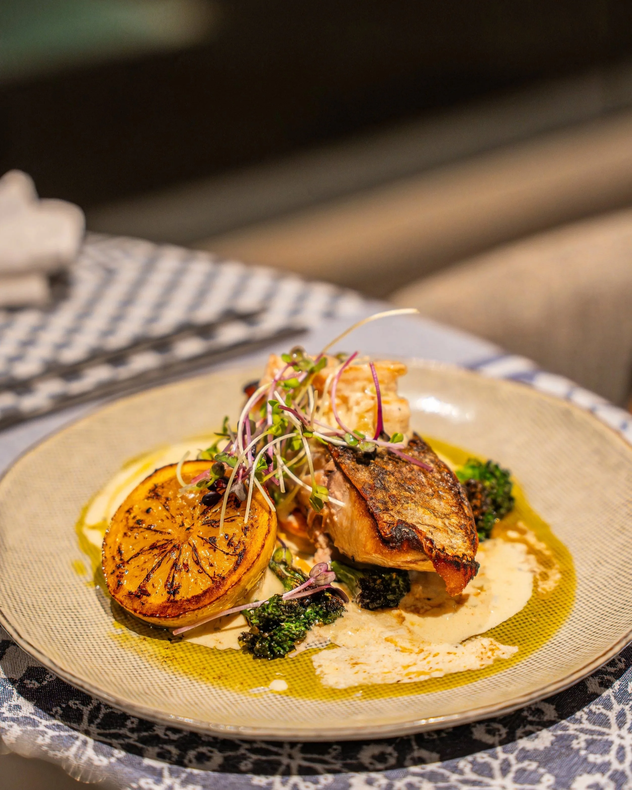 A plated dish with grilled seafood, lemon slices, microgreens, and a creamy sauce, served on a decorative yellow plate.