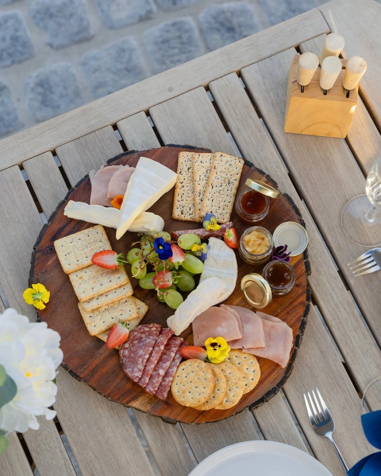 Charcuterie board with cheeses, meats, grapes, crackers, and jams on a wooden platter, set on an outdoor wooden table with a cobblestone background.