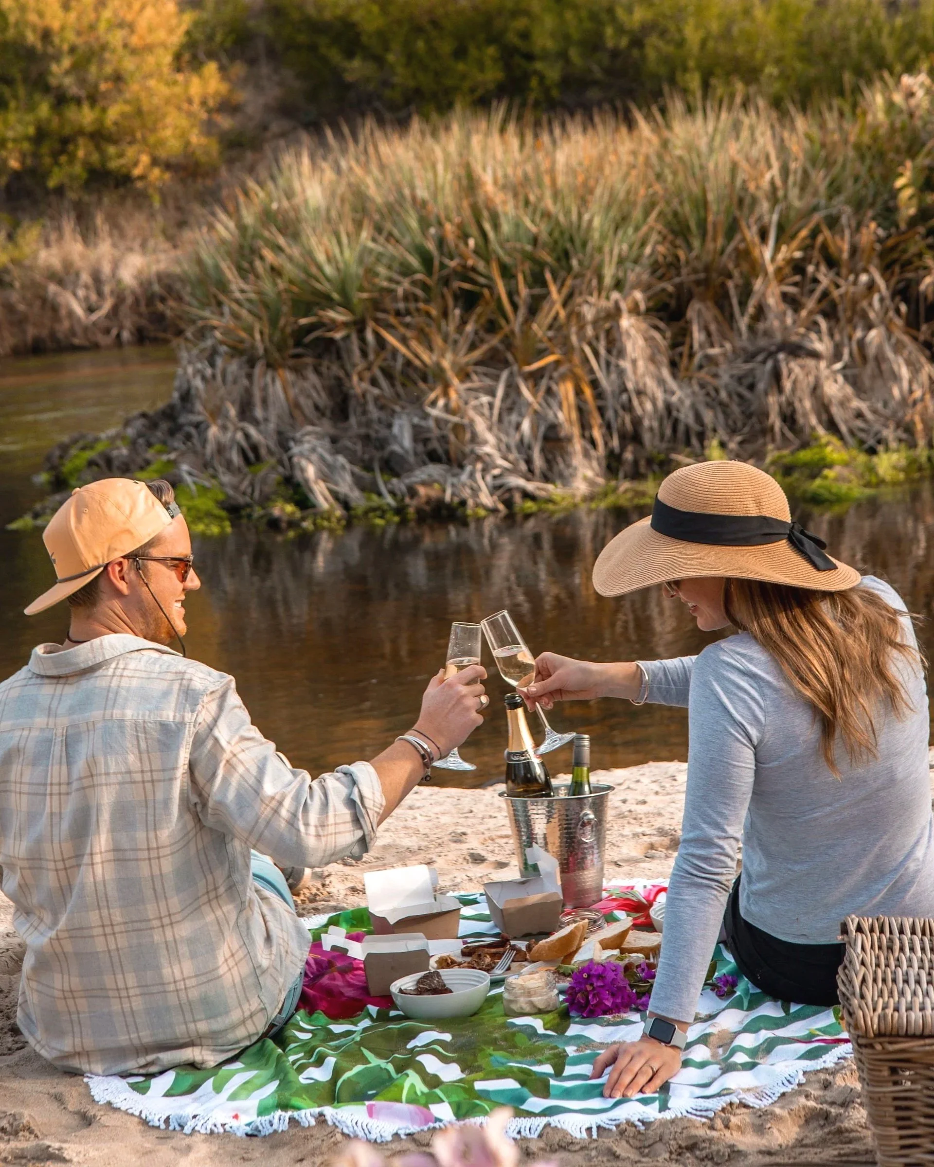 A man and woman having a picnic on a sandy riverbank, clinking champagne glasses, with food and a bouquet of flowers on a blanket.