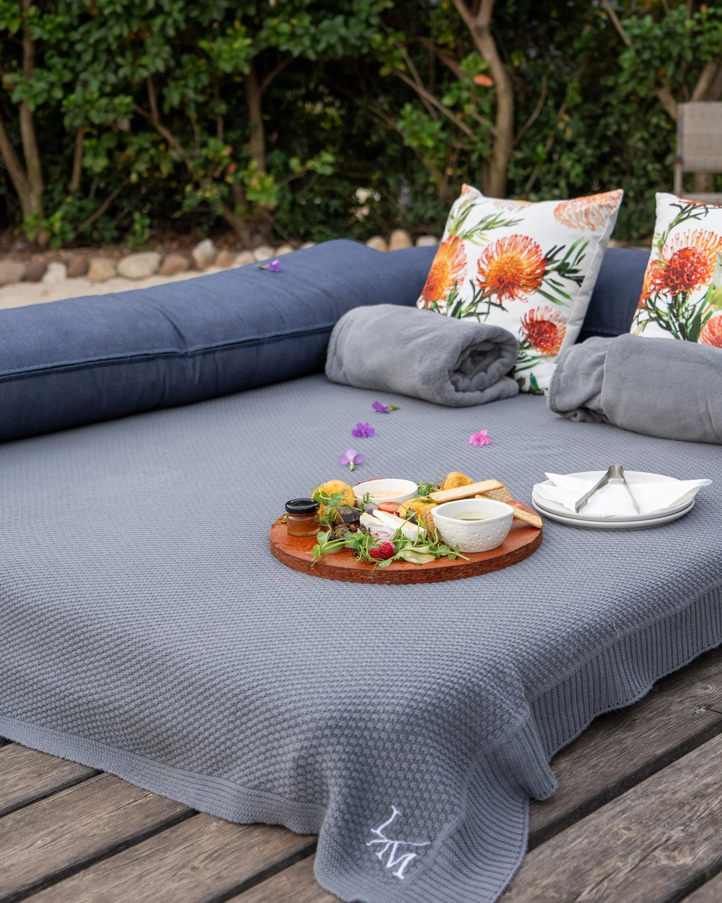 Outdoor lounge area with a large, textured gray blanket draped over a wooden surface, decorative pillows with floral patterns, a tray of snacks and drinks, and a background of lush green bushes.