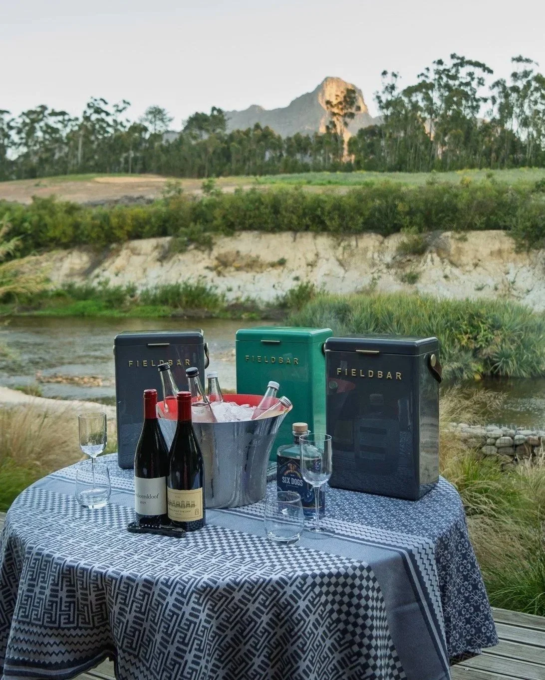 A table set for a outdoor gathering with wine bottles, glasses, and cooler boxes, overlooking a river and a mountain in the background.