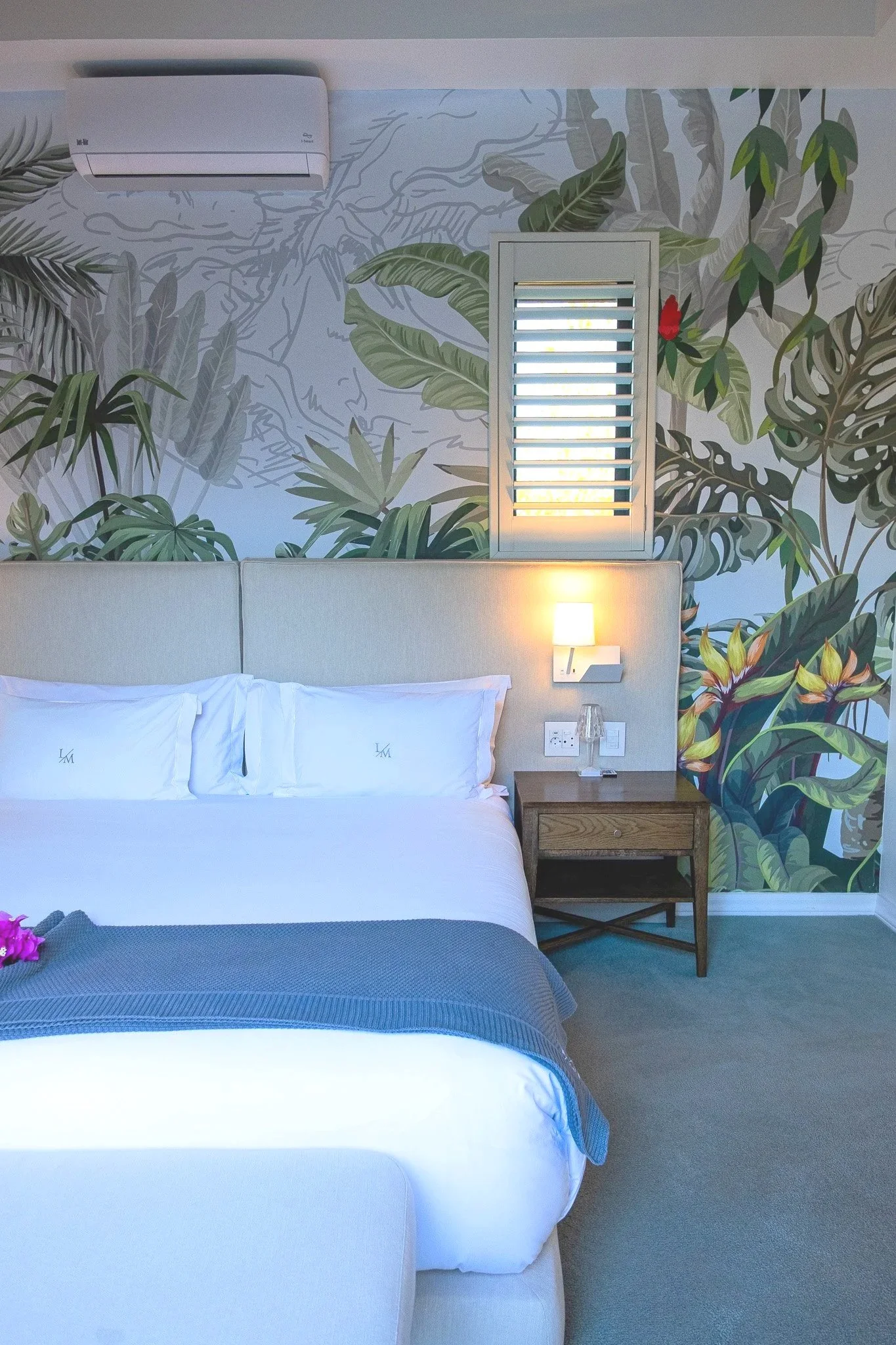 A hotel room with a white bed, a small wooden bedside table, a wall-mounted lamp, and a tropical jungle-themed mural on the wall featuring various green leaves and plants, with a window that has white shutters and a small air conditioning unit above.