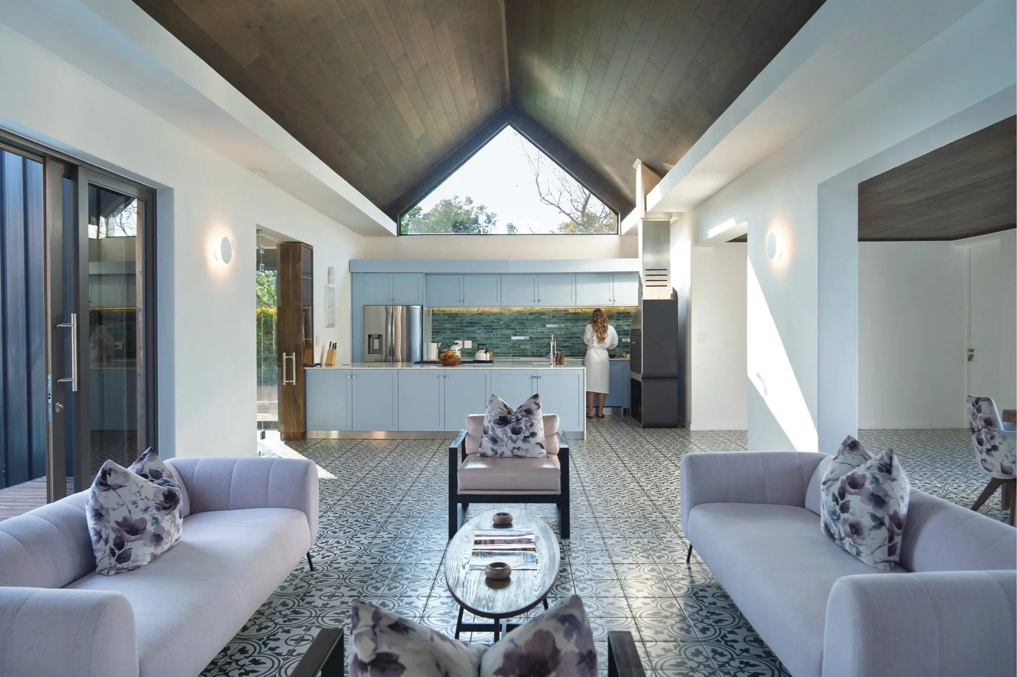 A modern living room with patterned tile floor, white sofas with floral pillows, and a view into a kitchen with blue cabinets and a vaulted ceiling with a large triangular window.