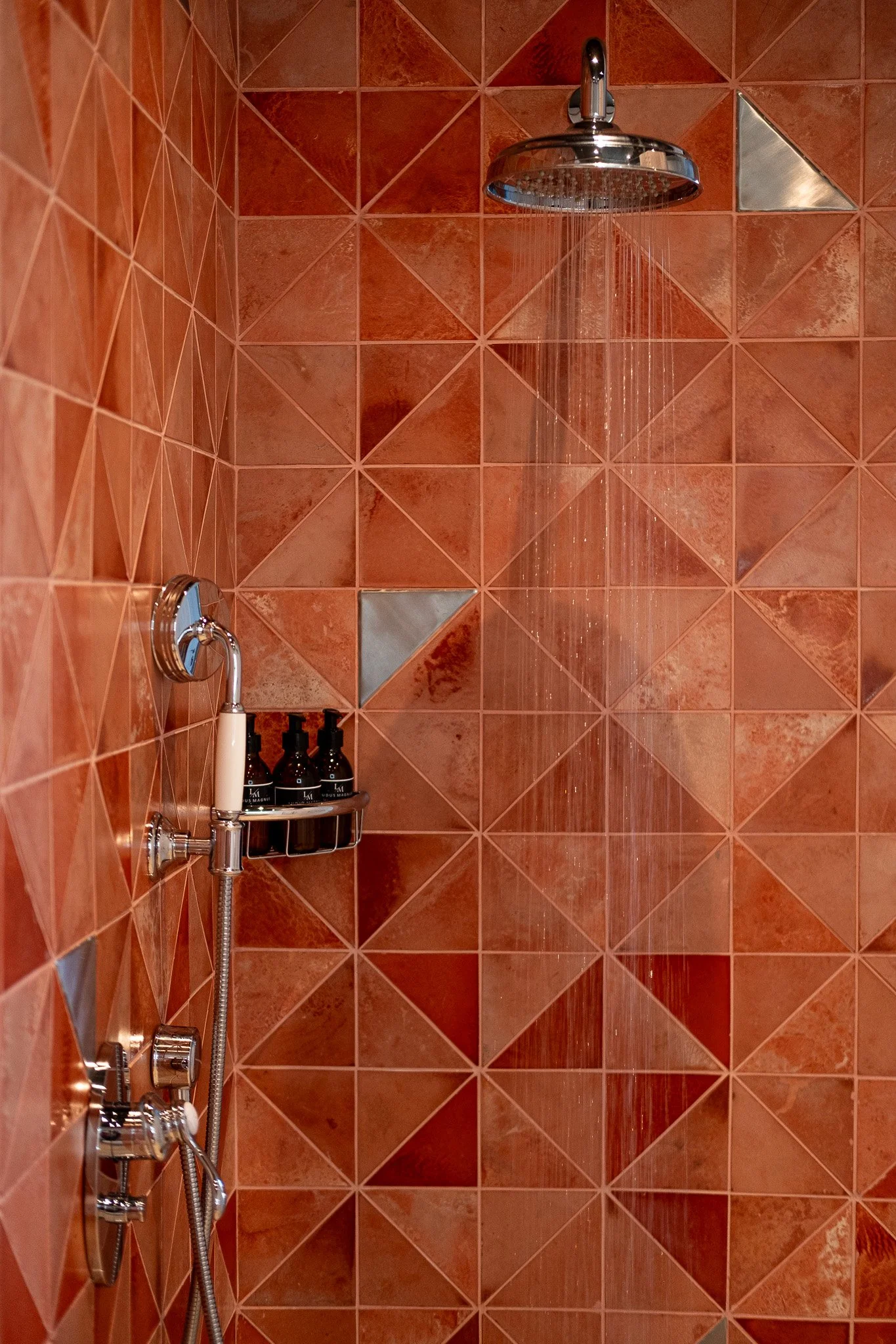 A shower with a chrome rainfall showerhead installed on a wall covered with red and orange geometric patterned tiles. A shelf holding three black bottles and a chrome faucet with a flexible hose are visible.