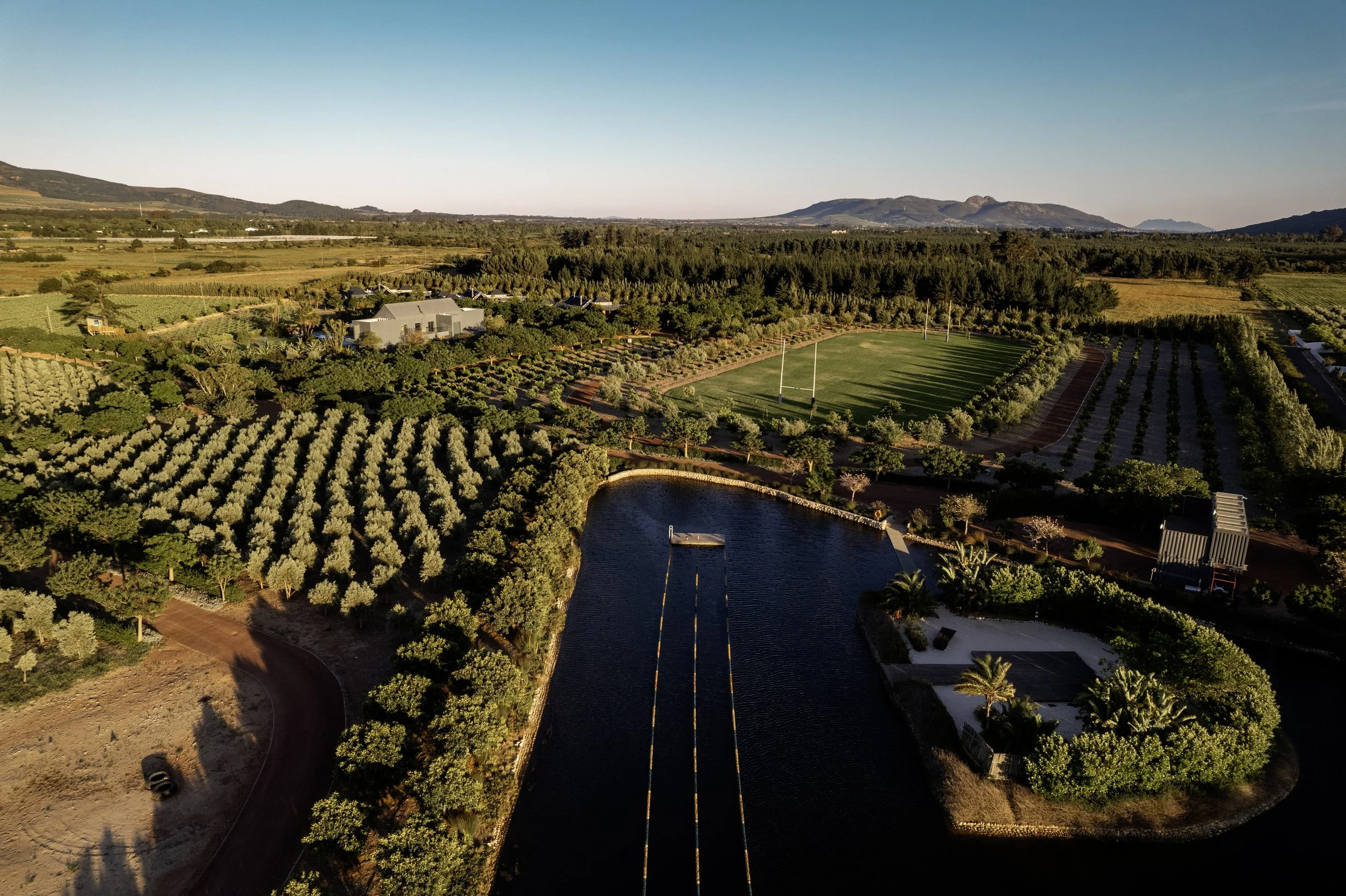 Aerial view of a scenic landscape with a pond, lush green fields, trees, and distant mountains under a clear blue sky.