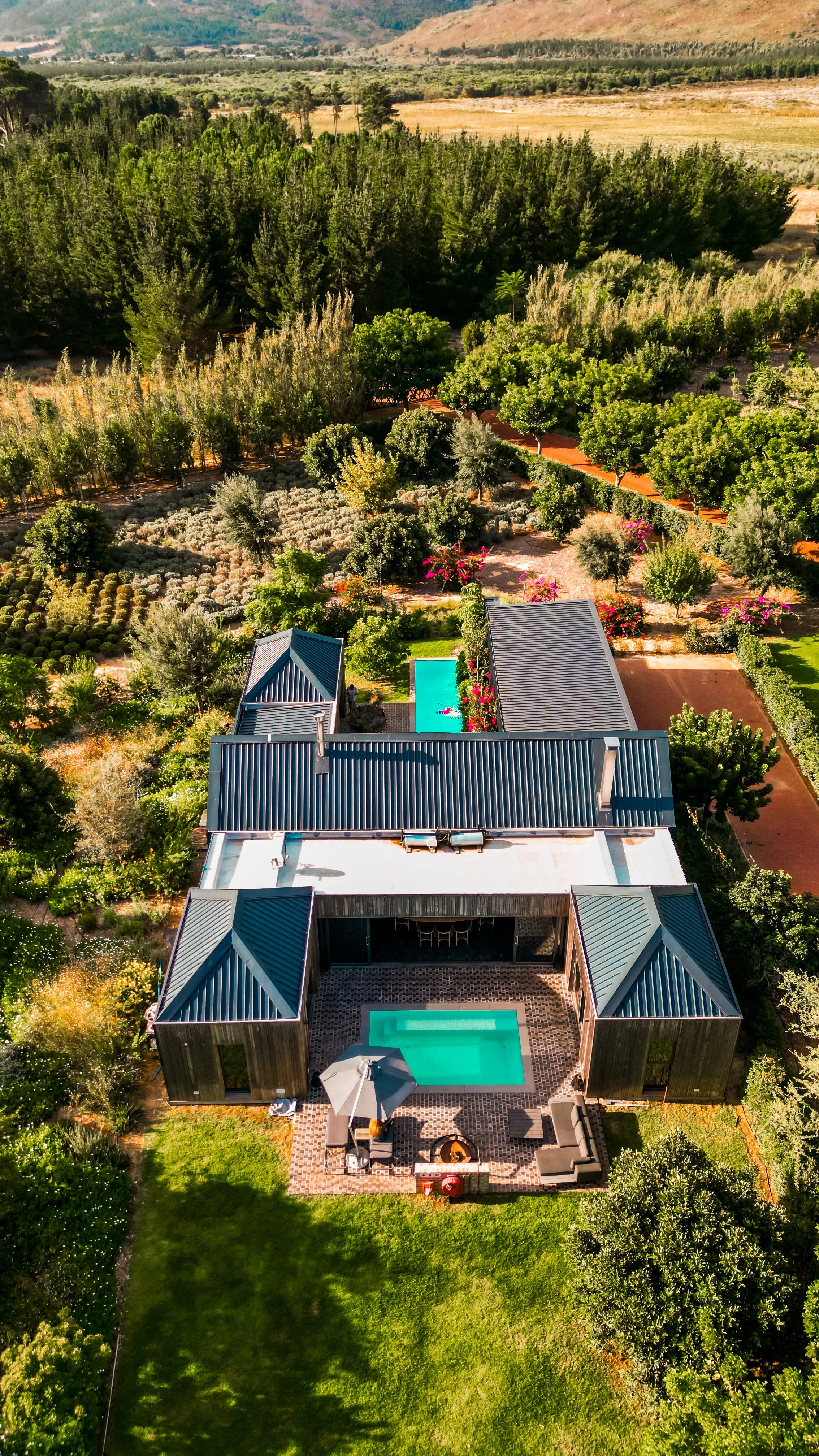 Aerial view of a modern house with a backyard swimming pool, patio furniture, and a large umbrella, surrounded by lush greenery, trees, and landscaped gardens, with fields and hills in the background.