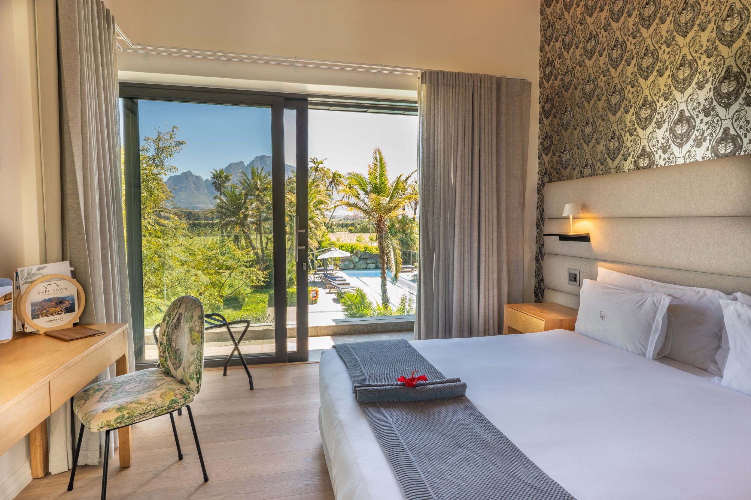 A bright hotel room with a view of tropical trees, mountains, and a swimming pool outside. The room has a bed with white linens, a small wooden nightstand, and a wall mounted lamp at Ludus Magnus 5 star Luxury Boutique Hotel and Villas