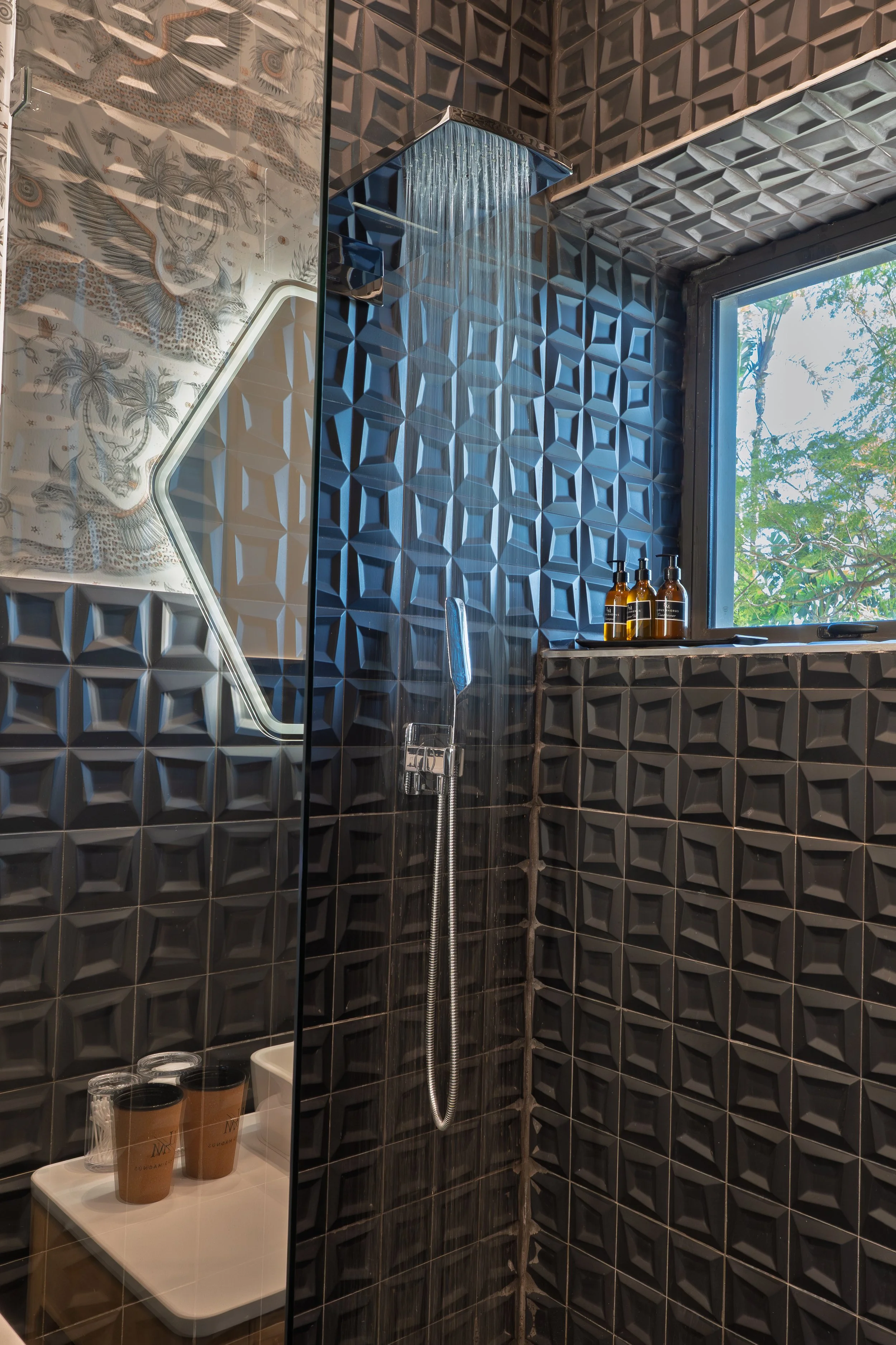 Bathroom shower with black geometric 3D tiles, a window showing trees outside, three bottles on the window ledge, a hexagonal mirror, and a small shelf with two coffee cups.