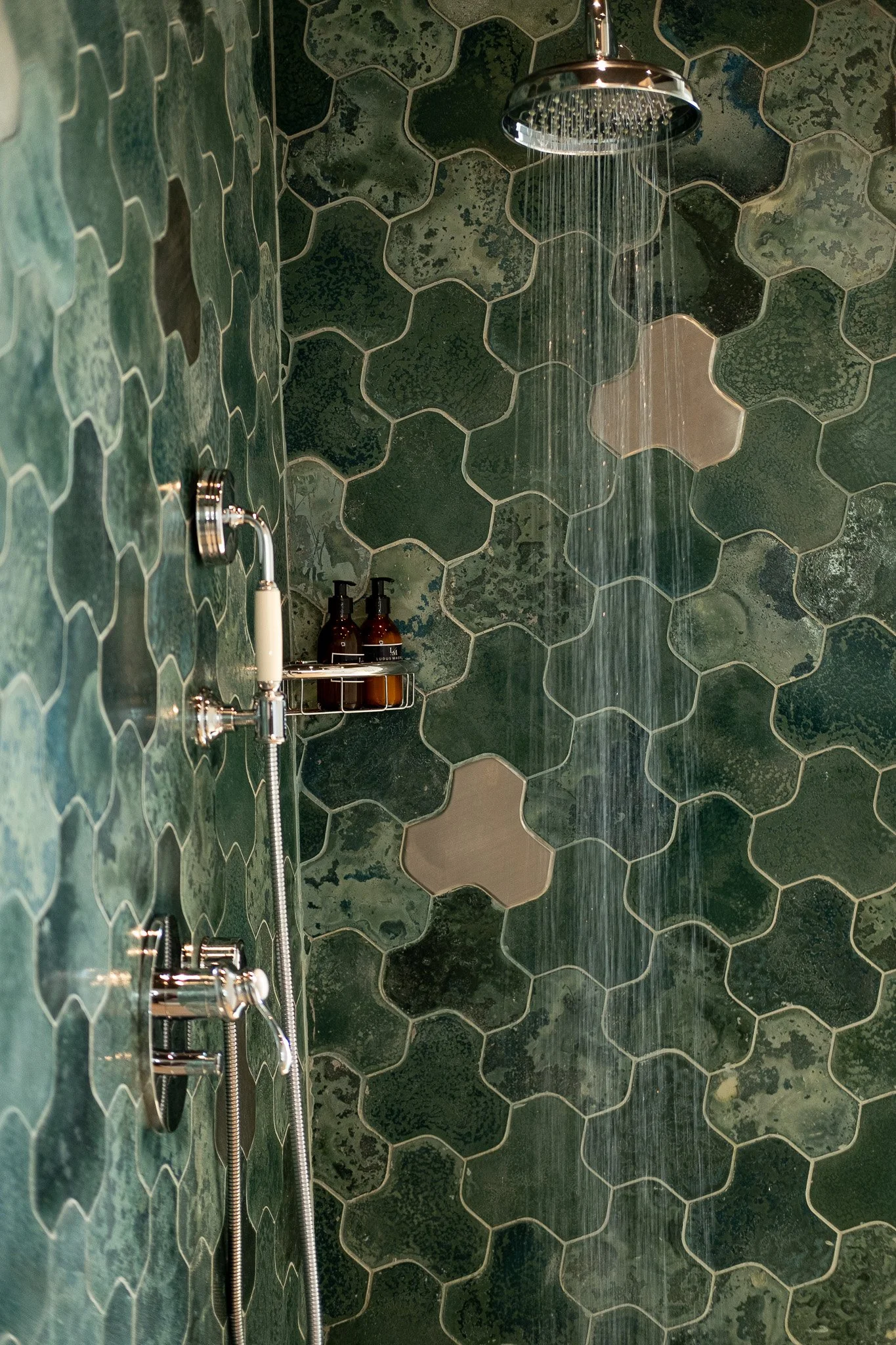 Green hexagonal tiled shower wall with a chrome showerhead, a handheld shower hose, and soap dispensers.