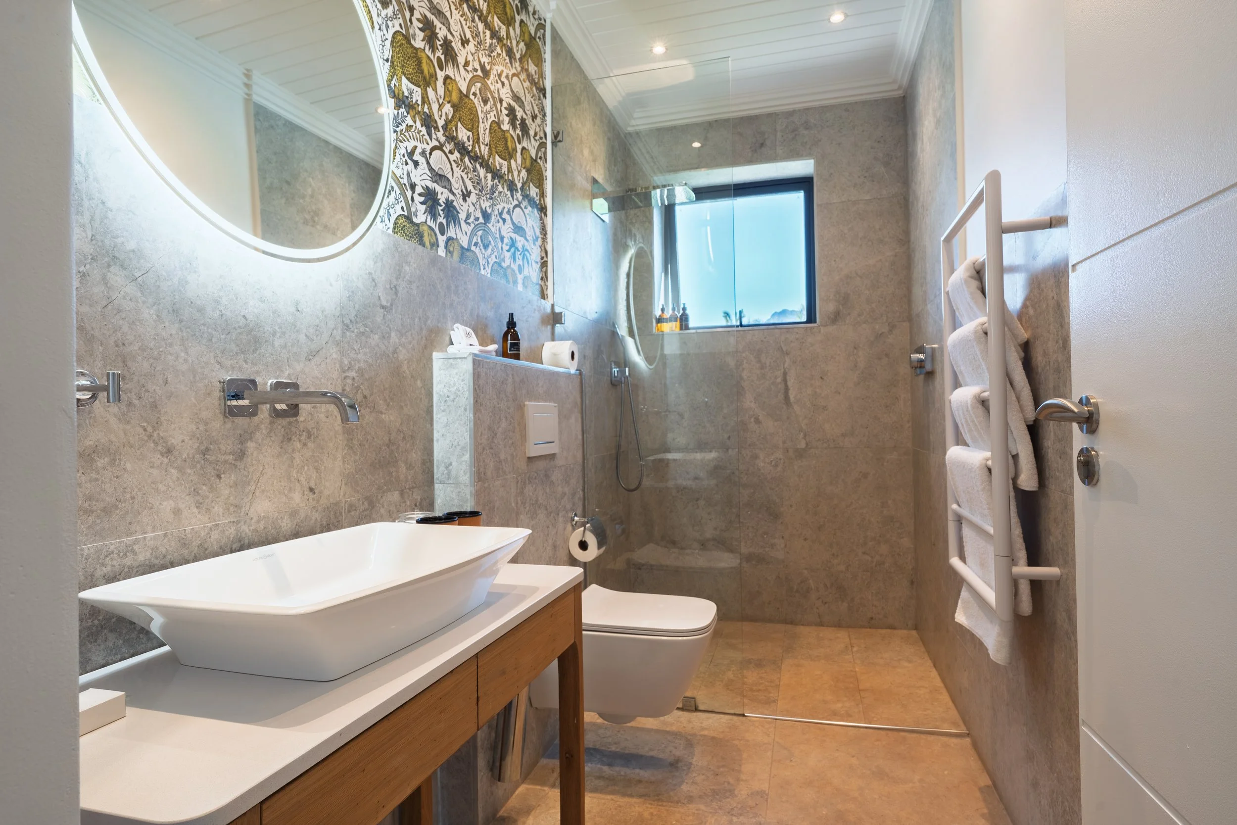 Modern bathroom with beige marble tiles, a freestanding bathtub, a vanity with a vessel sink, a wall-mounted mirror, a toilet, a heated towel rack with towels, a glass shower enclosure, a window with a view, and a stylish decorative tile accent.