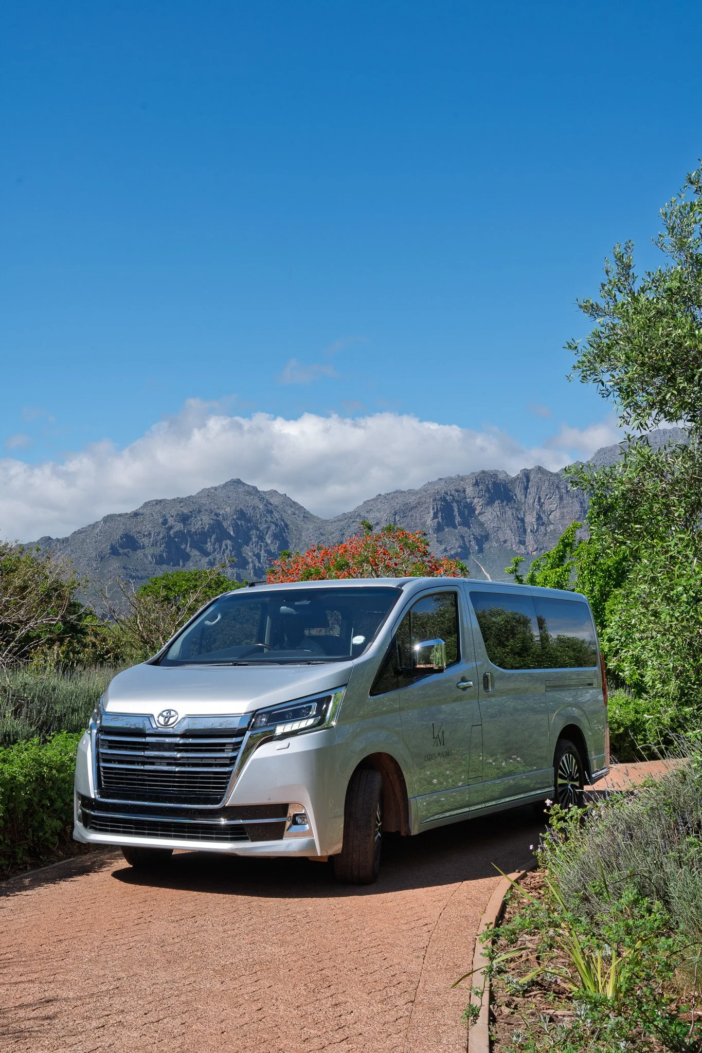A silver Toyota van parked on a dirt road, with green trees and mountains in the background under a blue sky with some clouds.
