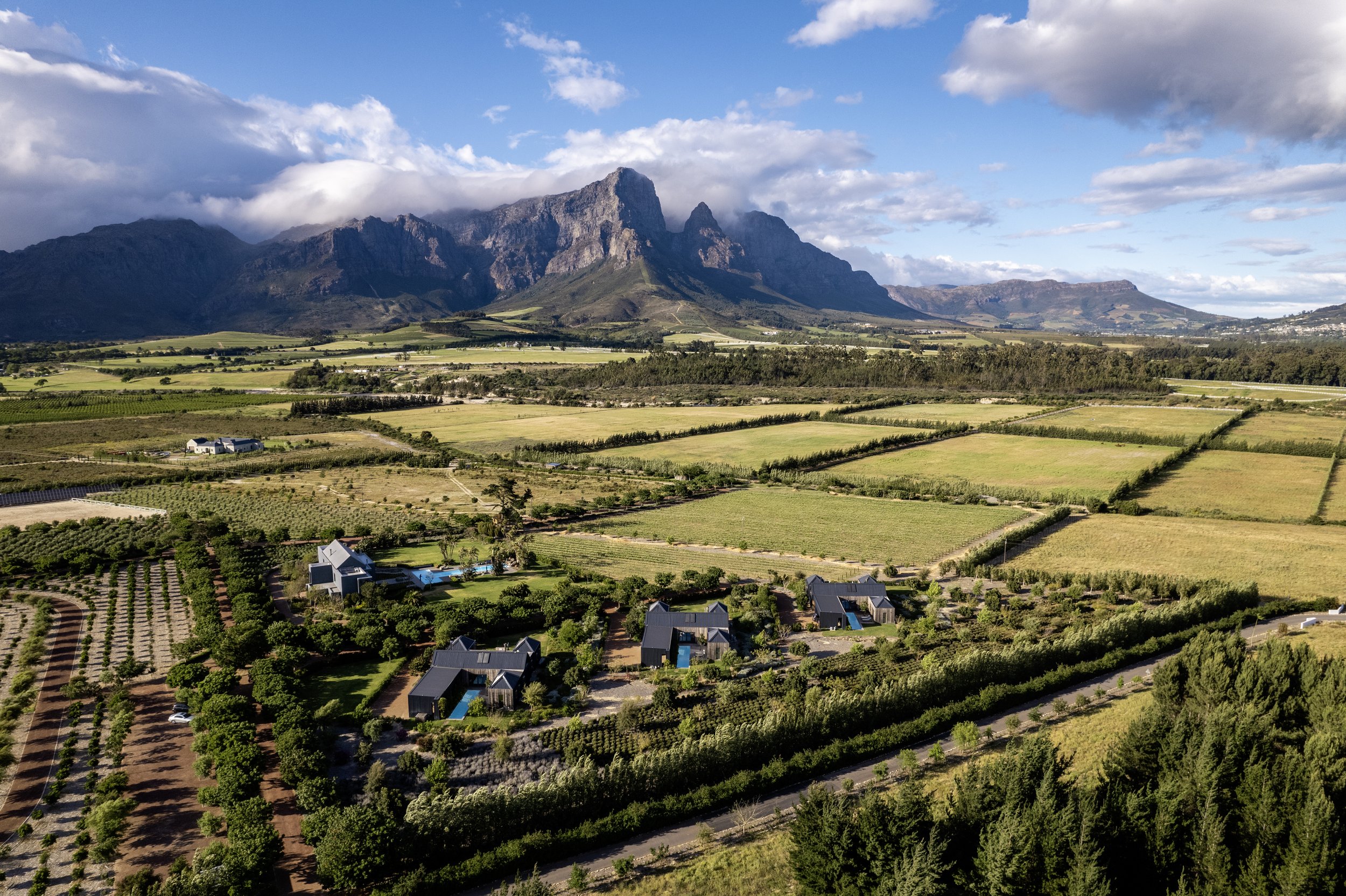 Aerial view of a rural landscape with green fields, small buildings, and trees with a mountain range in the background under a partly cloudy sky.