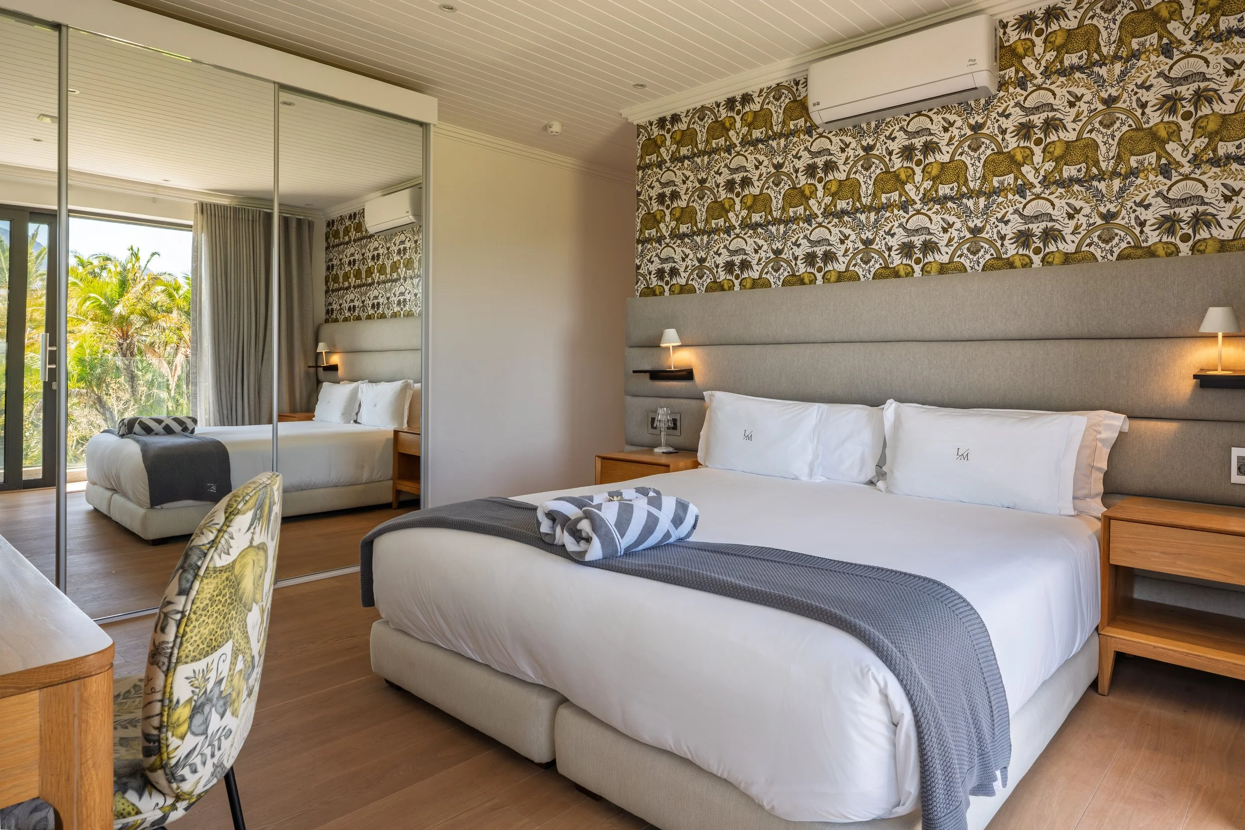 A hotel room with two beds, one in the foreground with white linens and a gray throw, and one in the background with similar bedding. The room has a large window with curtains, mirrored closet doors, a wooden nightstand, and tropical-themed wallpaper