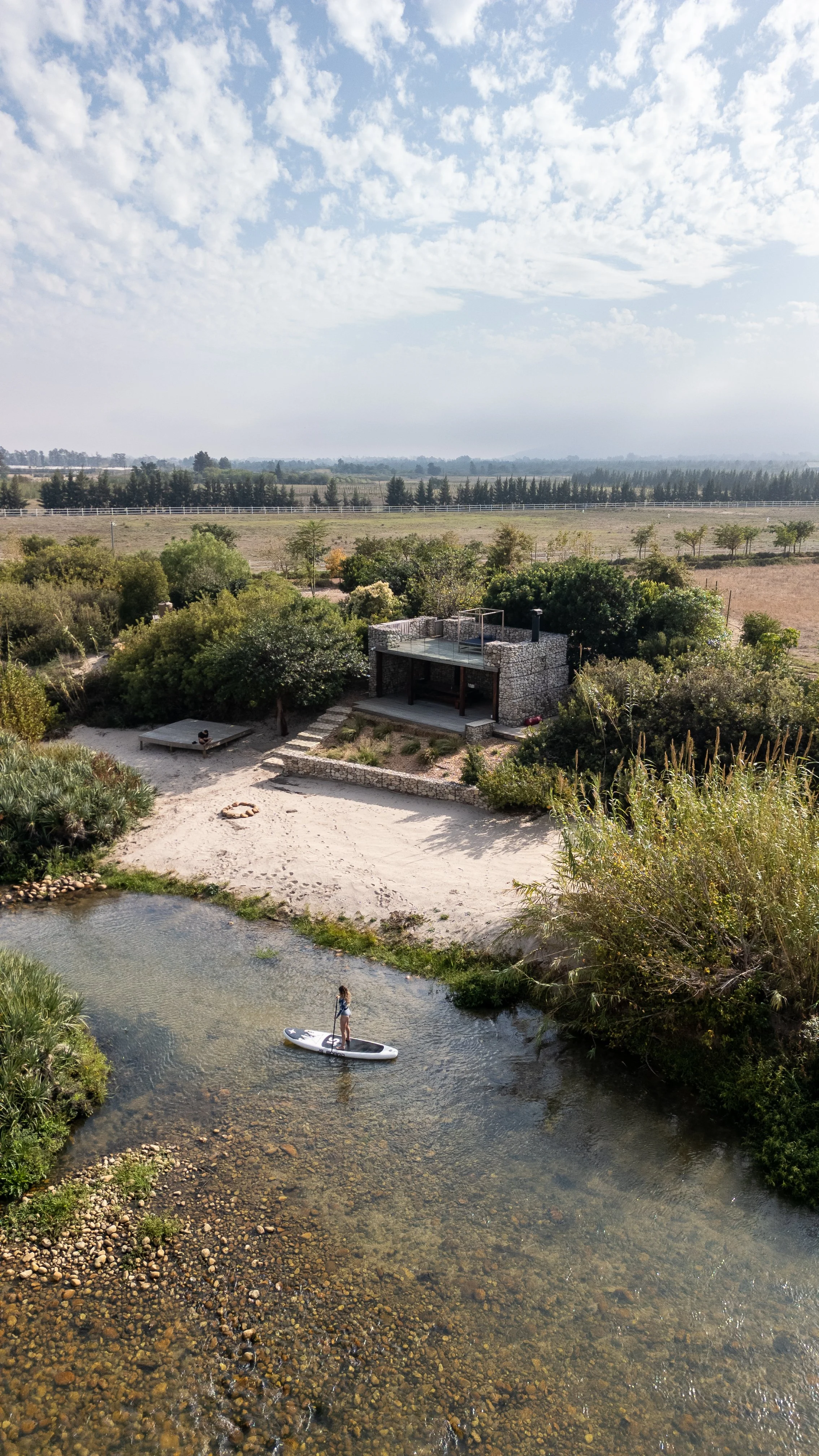 A woman paddleboarding in a river with lush trees and greenery, and a modern stone house on the bank under a partly cloudy sky.